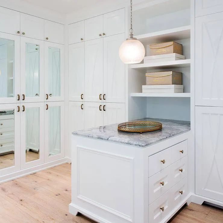 White walk-in closet with marble island, shelves, pendant light, and mirrored doors, on light wood floor.