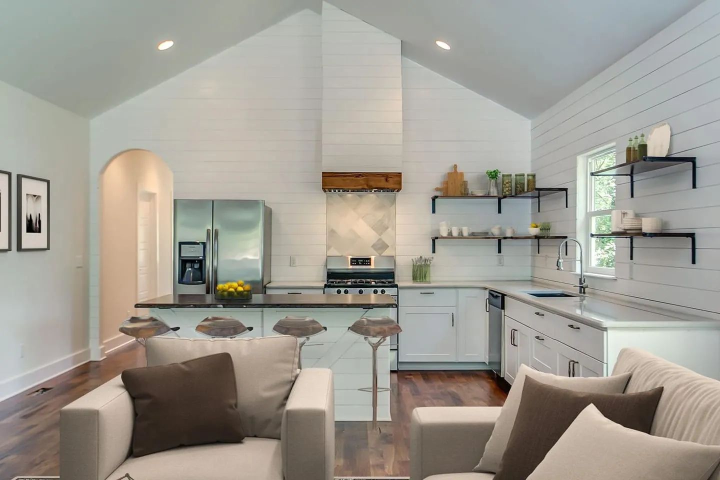 Cozy white kitchen with vaulted ceiling, stainless steel appliances, open shelving, and seating area with sofas.