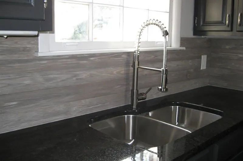 Kitchen sink with a chrome faucet, dark countertop, and gray wood-look backsplash.
