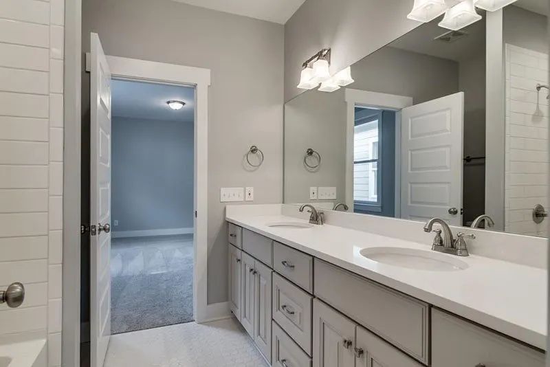 Bathroom with double sinks, gray cabinets, white countertop, and doorway to a bedroom.