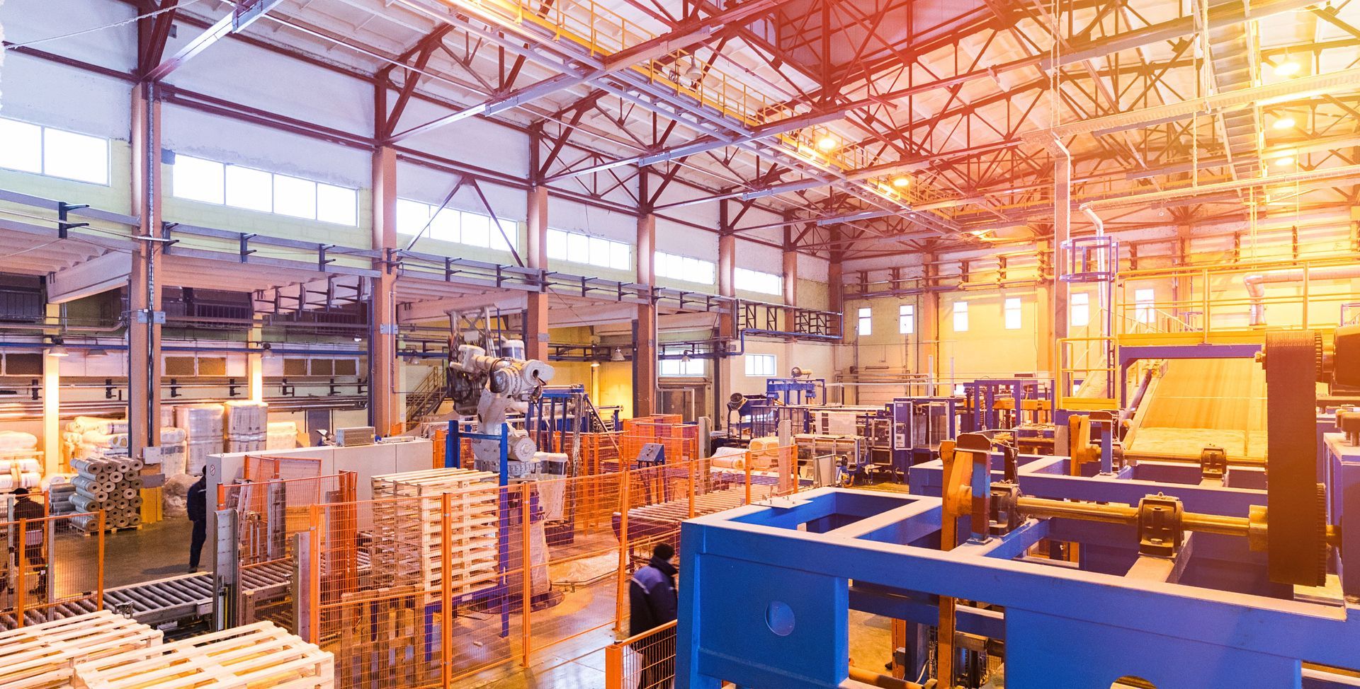 Factory interior with machinery, workers, and wooden pallets, lit by overhead lights.