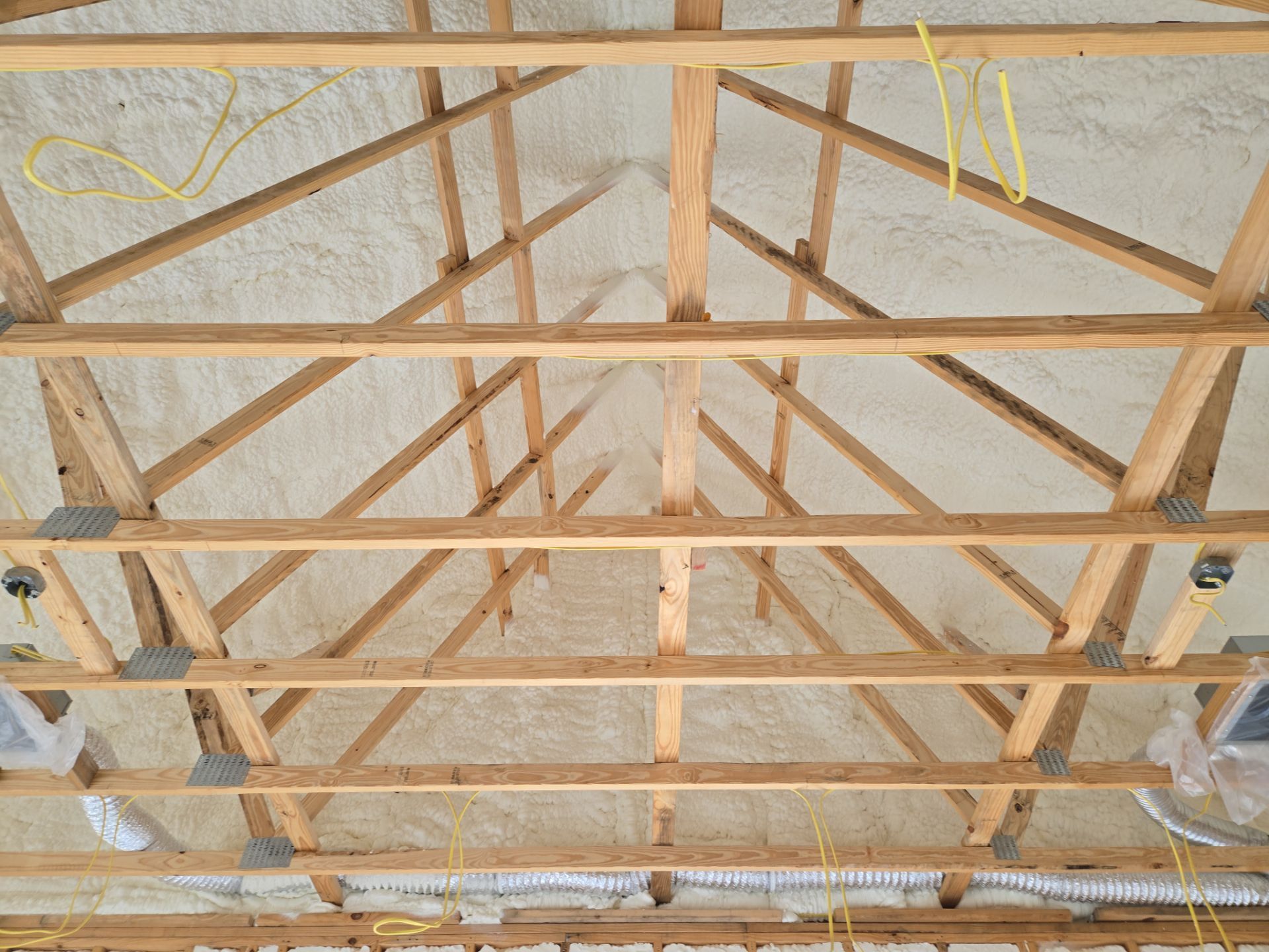 Wooden roof trusses with white spray foam insulation, visible wiring, and ductwork.
