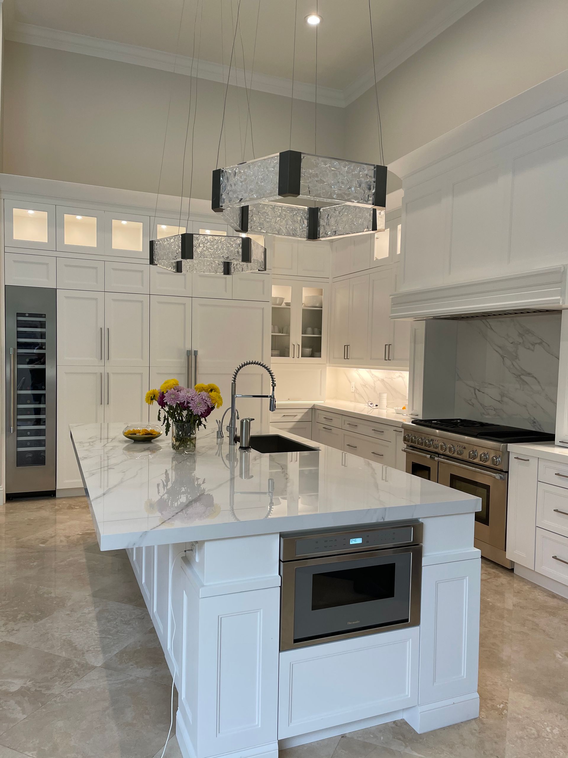 Bright, modern white kitchen with a large marble-topped island, stainless steel appliances, and two hanging chandeliers.