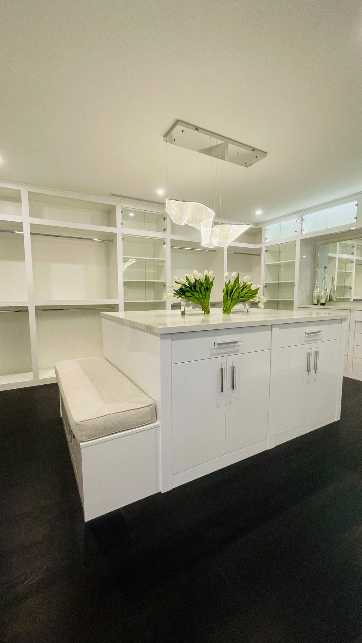 A white walk-in closet featuring a central island with a built-in bench, white cabinetry, and dark flooring.