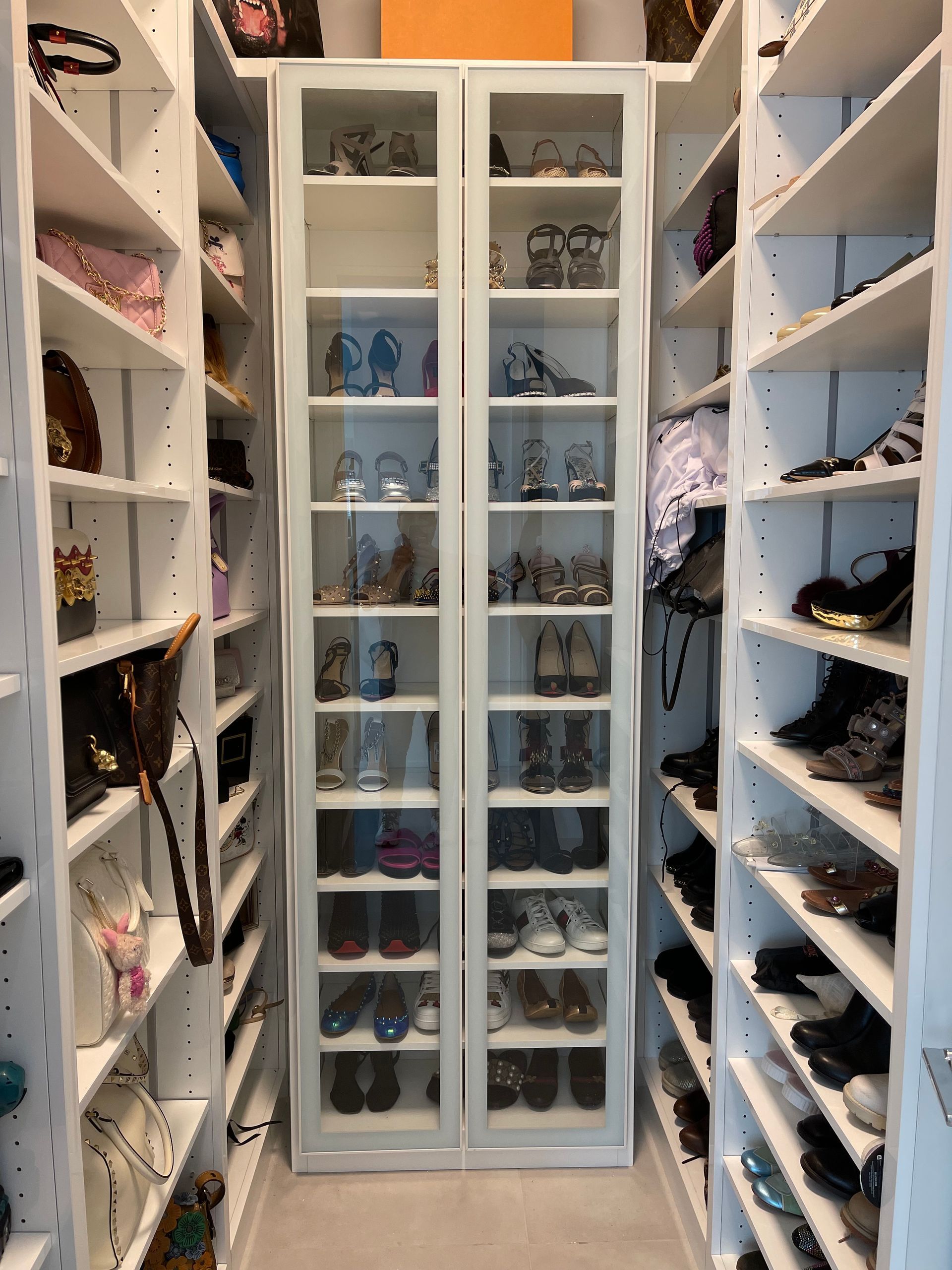 A white walk-in closet with open side shelves and a central glass-fronted cabinet displaying numerous pairs of shoes.