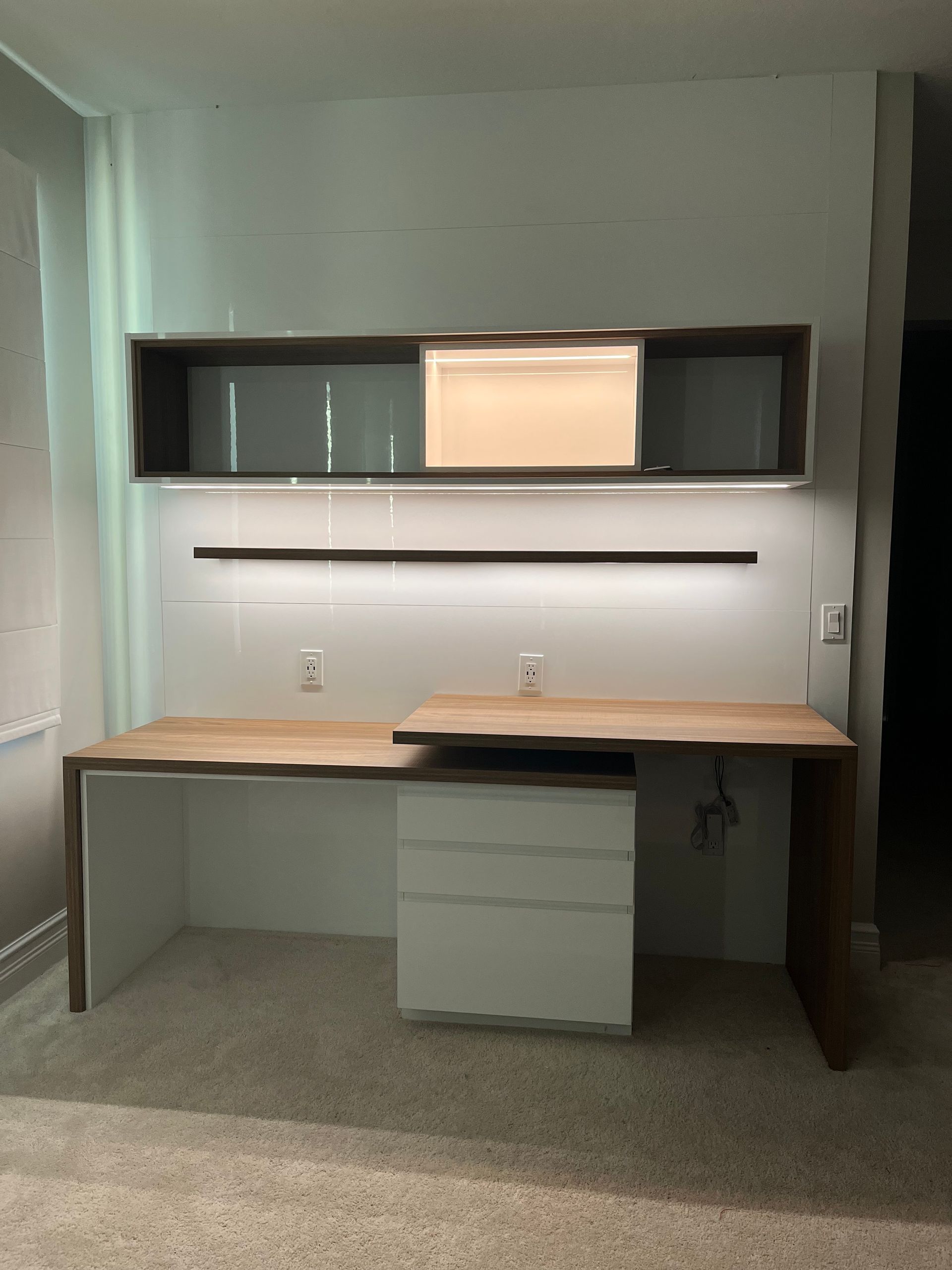 A custom floating desk setup with a wooden top, white drawers, and an illuminated wall-mounted shelf above on white wall.