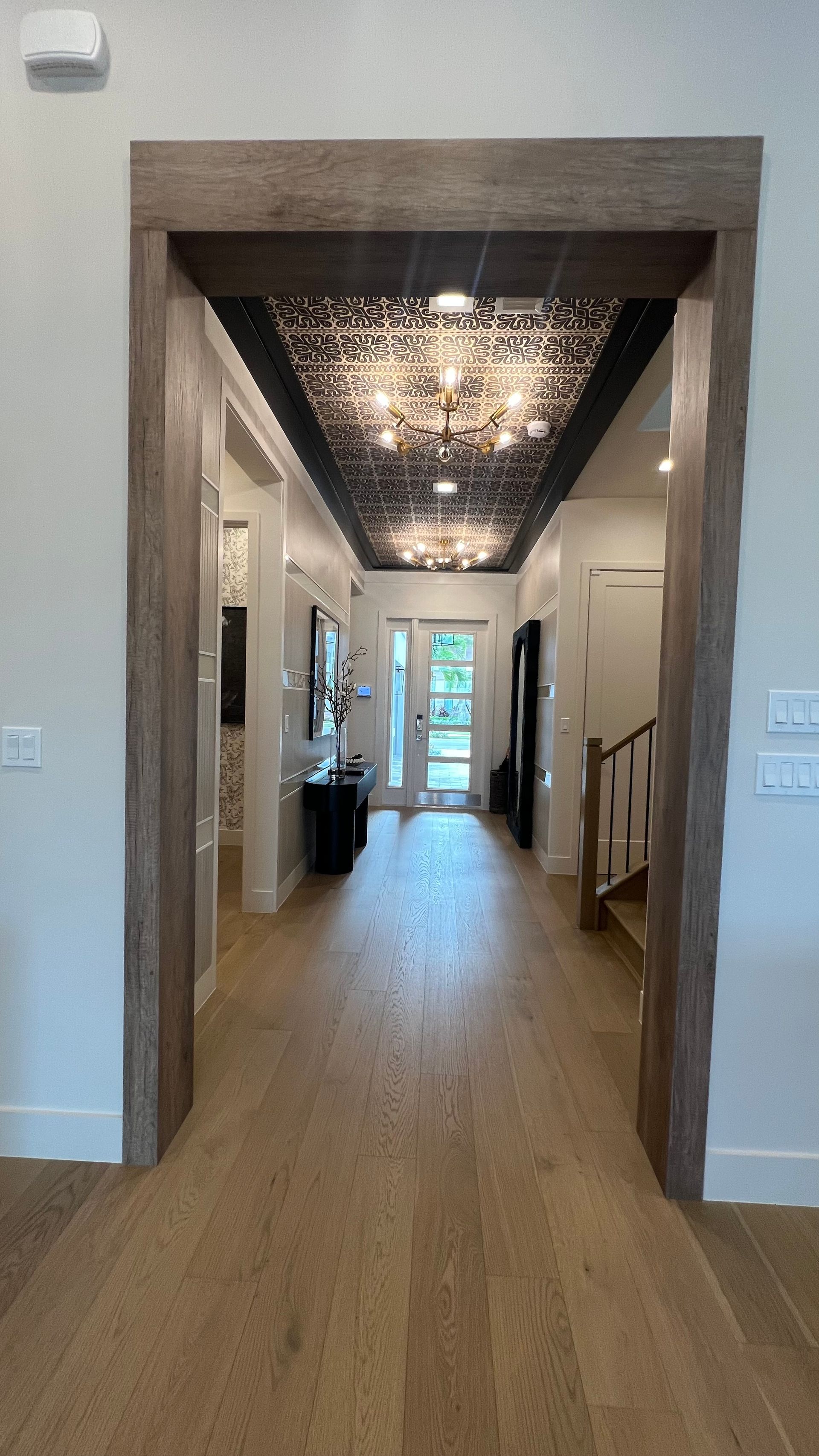A hallway view framed by a rustic wooden archway, featuring light wood floors and an ornate, dark ceiling with chandeliers.