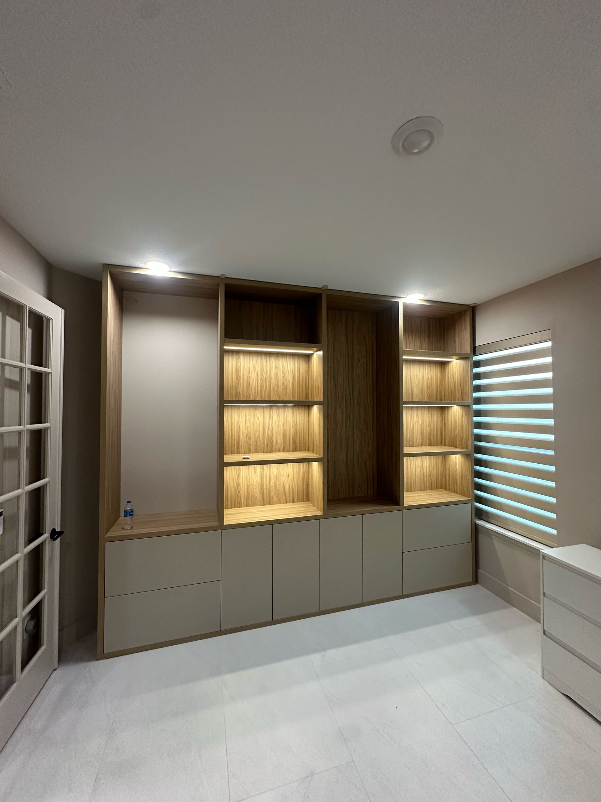 Built-in wooden shelving unit with integrated lighting and base cabinets against a neutral wall in a bright room.