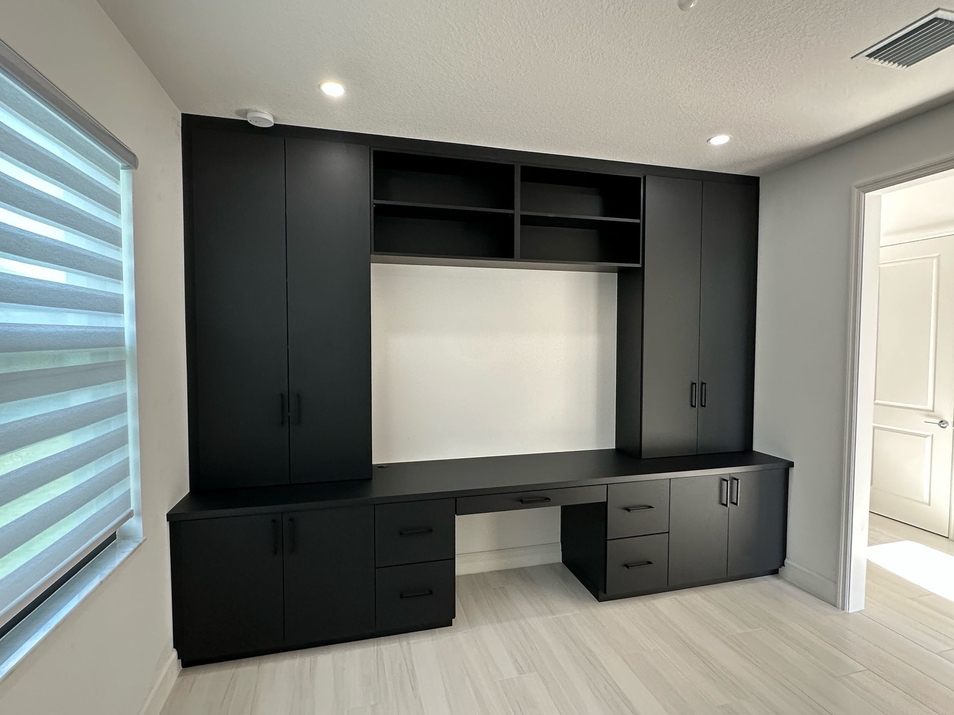 Matte black built-in office cabinetry with storage towers, shelves, and a central desk in a bright, modern room.