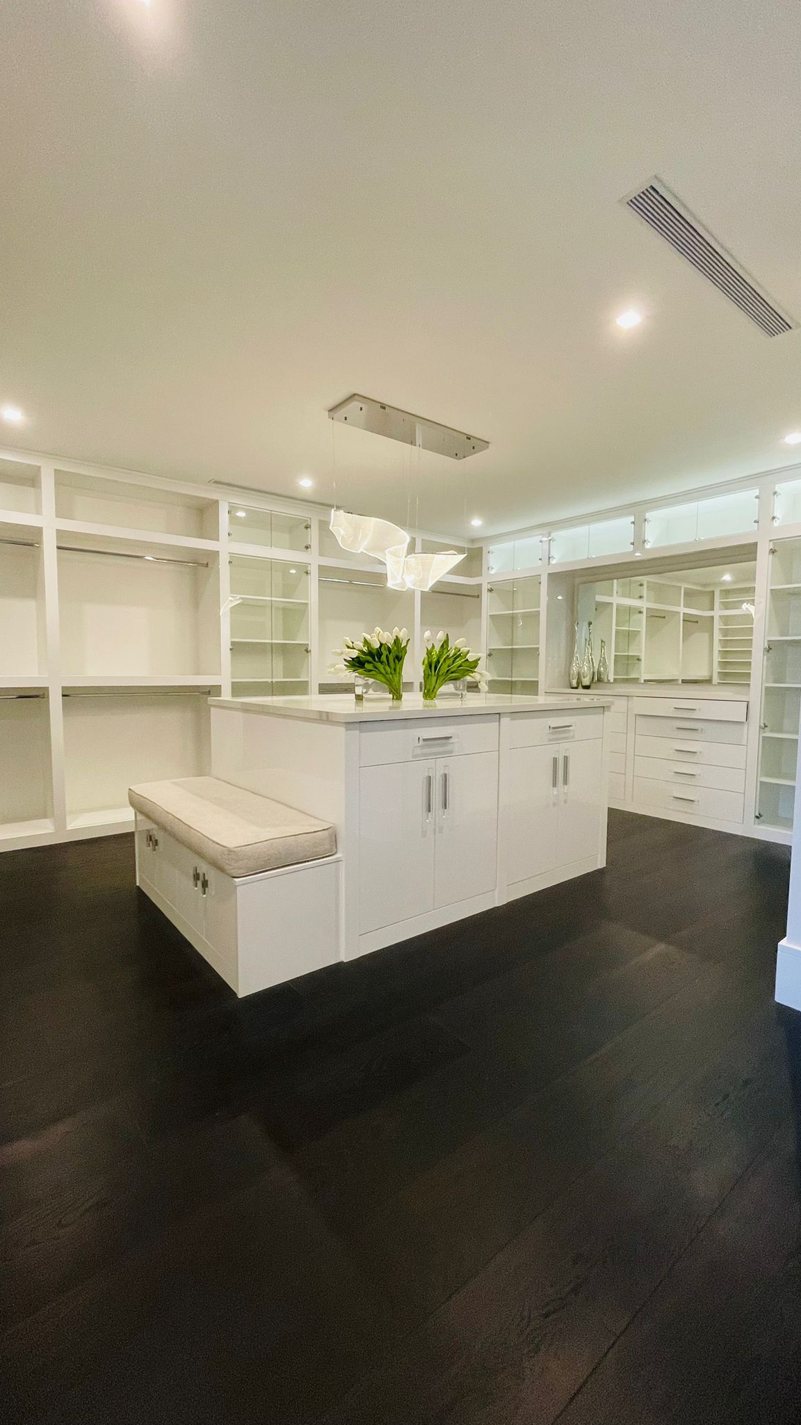 A spacious, modern walk-in closet with white cabinets, a central island with bench seating, and dark floors.