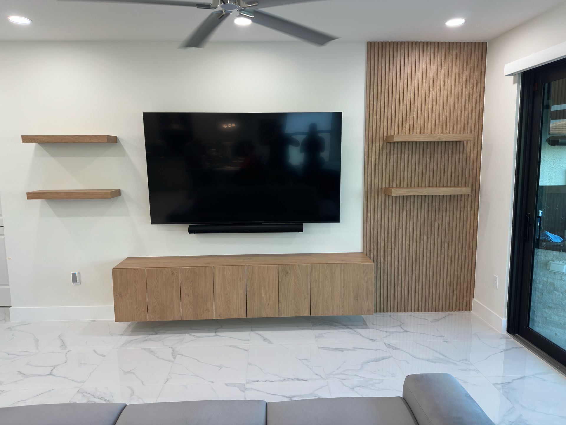 A wall-mounted TV over a floating light-wood console, flanked by floating shelves and a wood-slat accent wall.