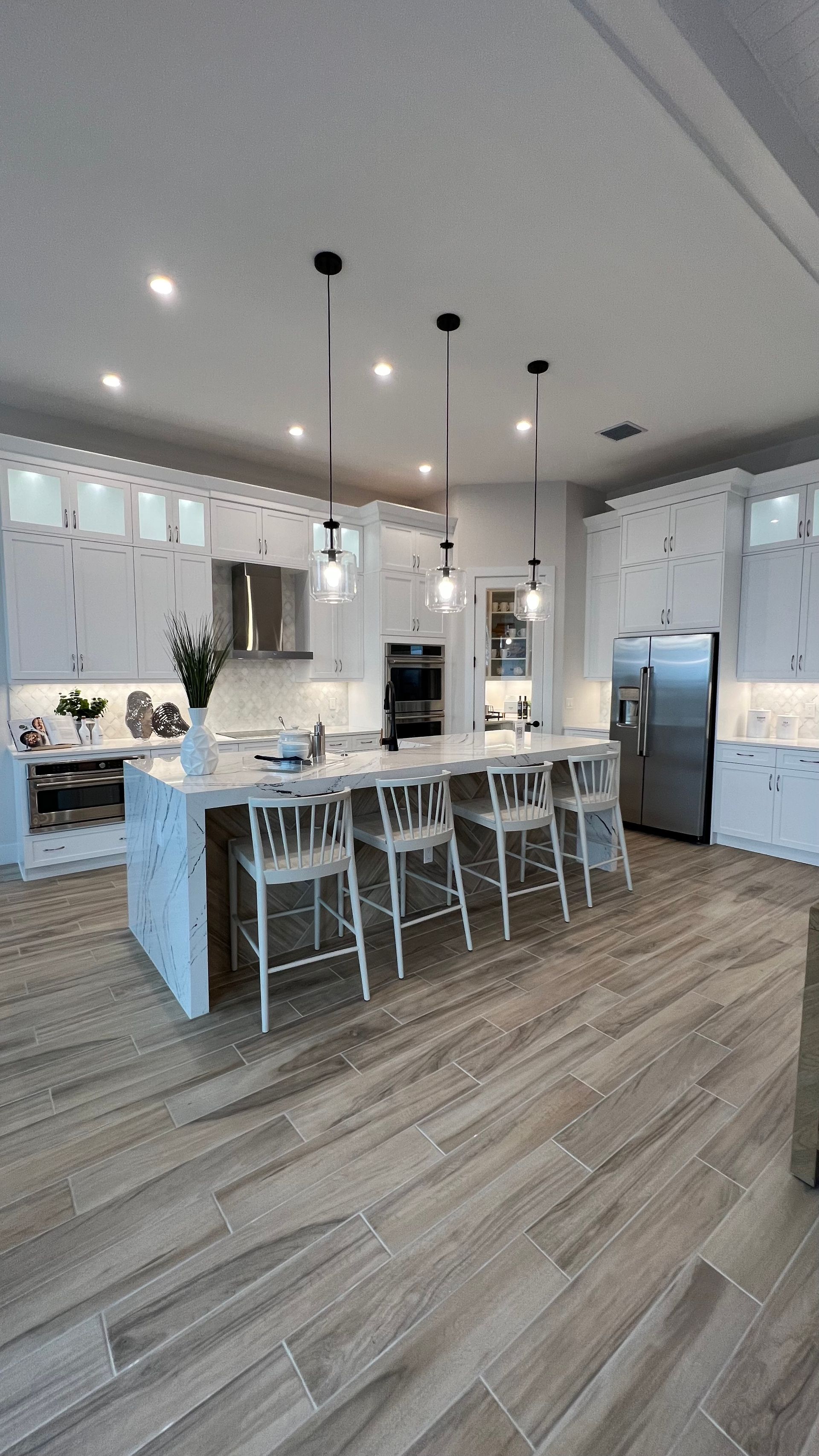 A modern, well-lit kitchen with white cabinetry, a large marble-topped island with four stools, and wood-look tile floors.