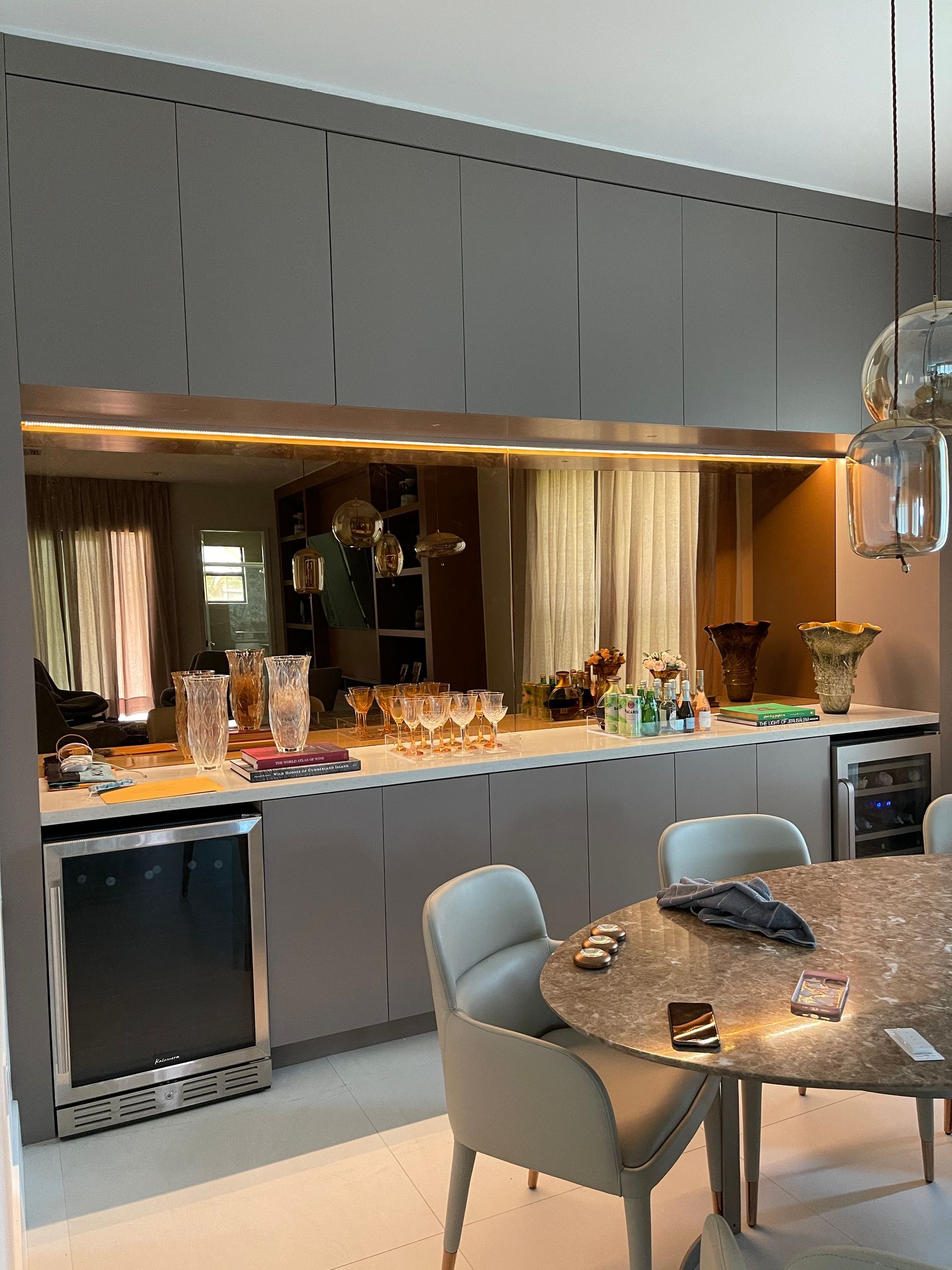 A sleek, gray home bar with a mirrored backsplash, a wine fridge, and a circular table with chairs in a dining room.
