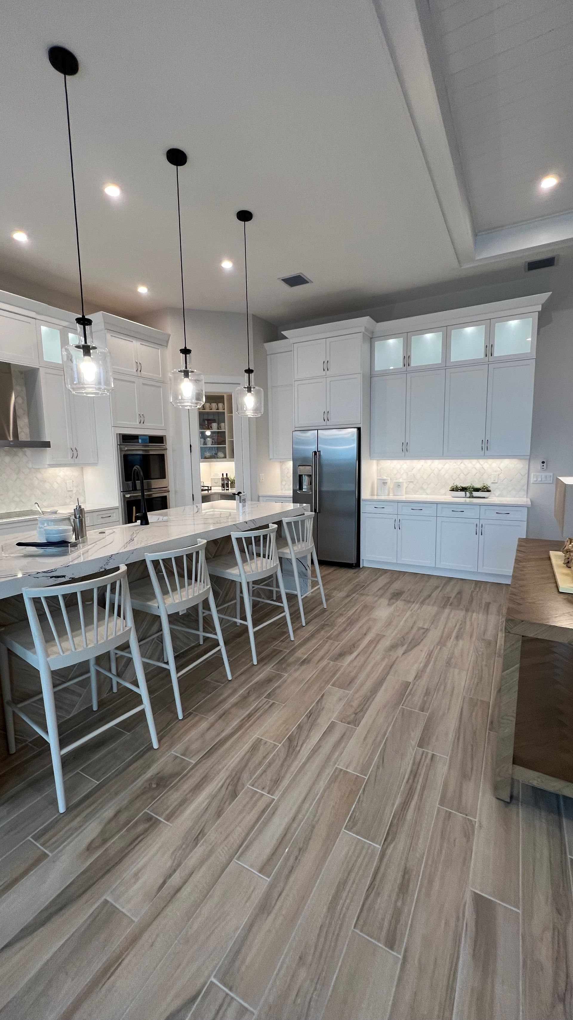 A modern kitchen with white cabinetry, light wood floors, a central island with four bar stools, and three pendant lights.