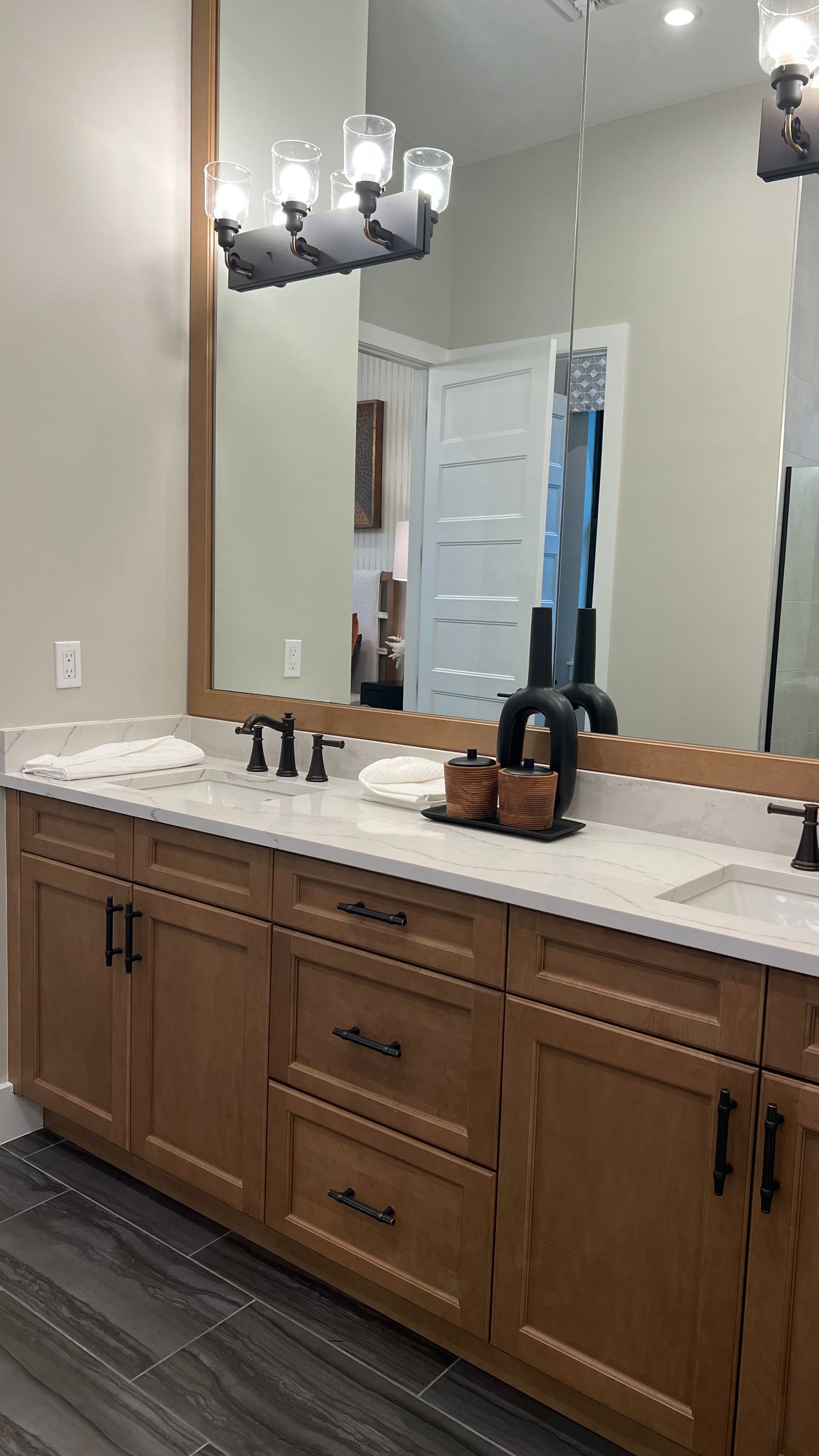 A modern bathroom vanity with wood cabinets, white countertops, a large mirror, and black hardware on a dark tiled floor.