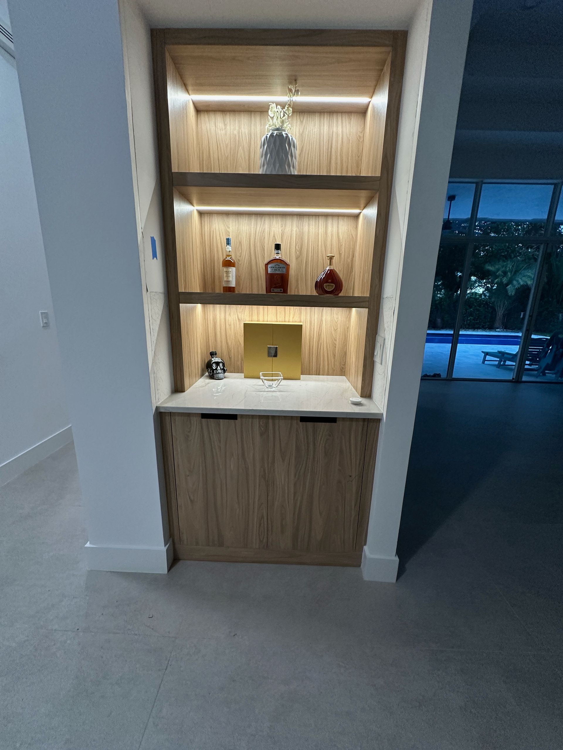 A built-in wooden bar nook with recessed LED shelving, holding a few bottles and a small gold box above a storage cabinet.