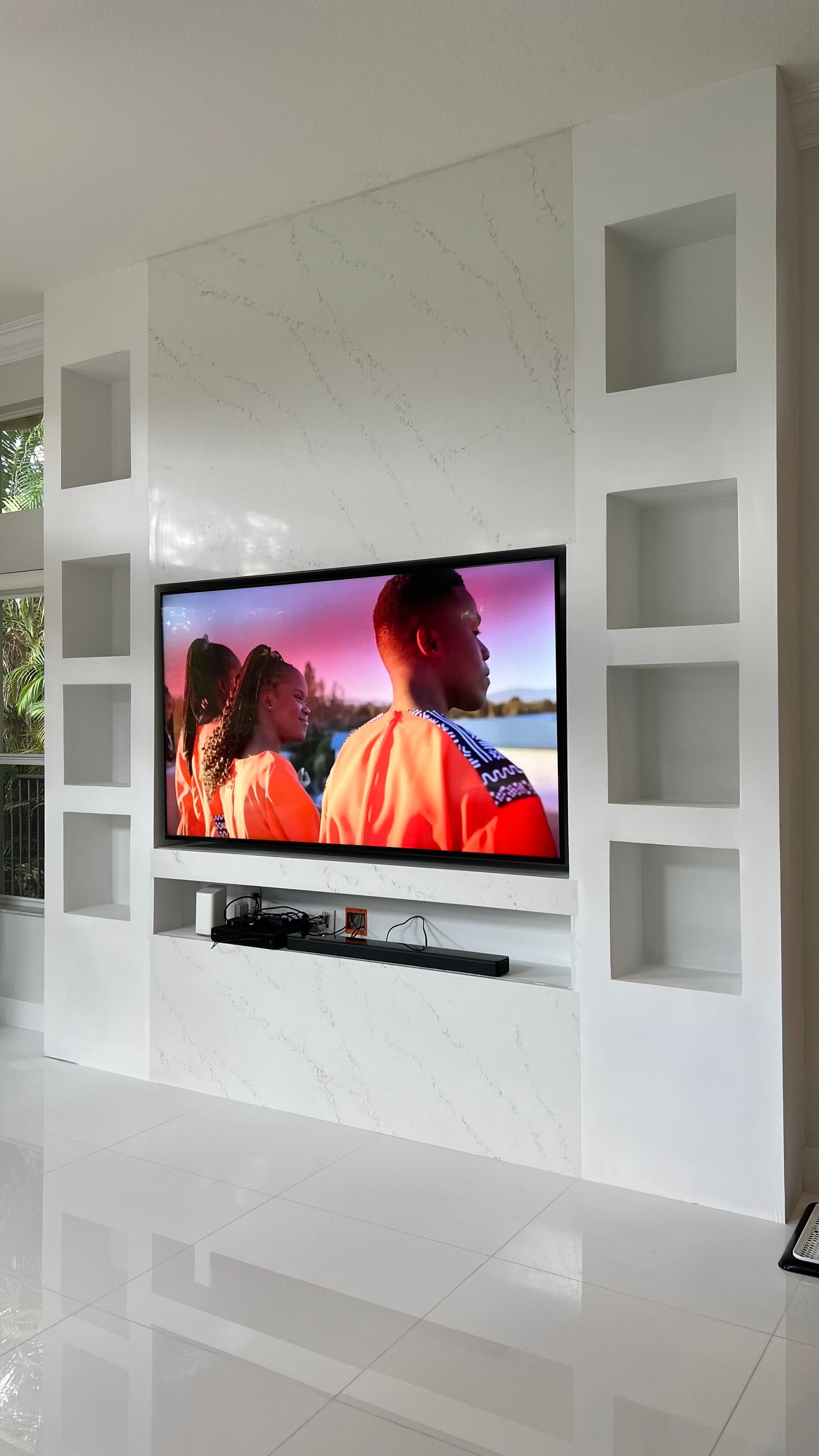 A wall-mounted TV on a white marble accent wall, flanked by white built-in shelving units in a bright, modern room.