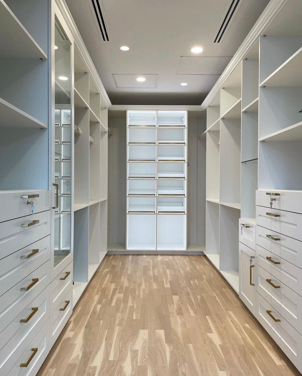 A modern walk-in closet with white shelving, numerous drawers, a central illuminated display unit, and light wood flooring.
