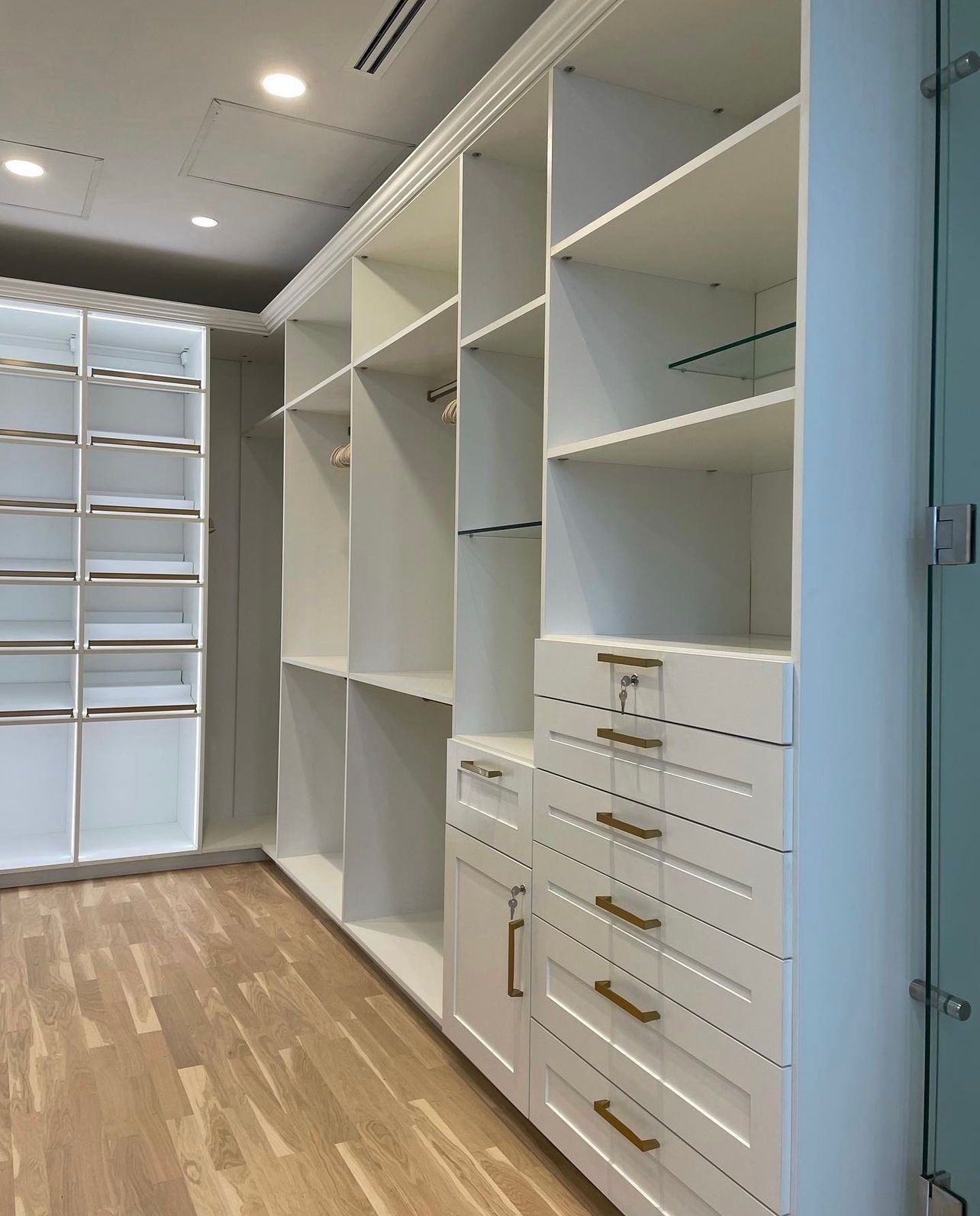 White custom walk-in closet shelving with gold hardware and integrated lighting, set against a light wood floor.