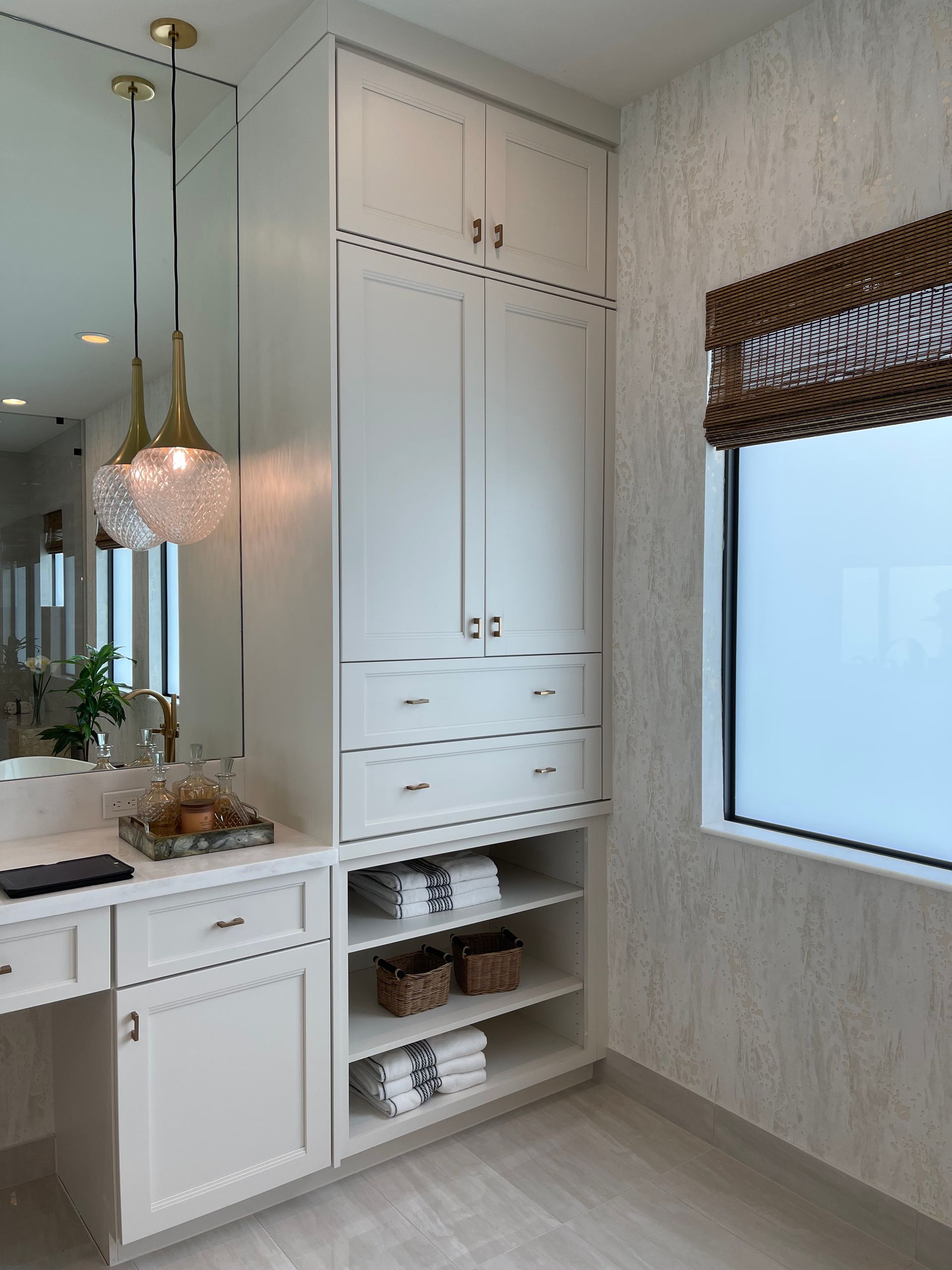 A bathroom vanity with white cabinets, drawers, open shelving, gold pendant lights, and a window with a woven shade.