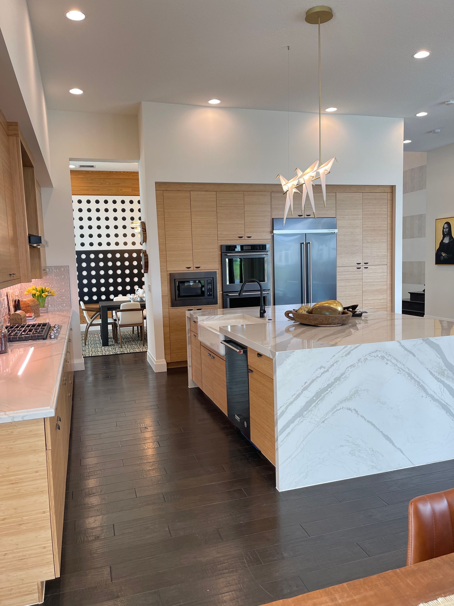 A modern kitchen with light wood cabinets, white marble countertops, a central island, and an open dining area beyond.
