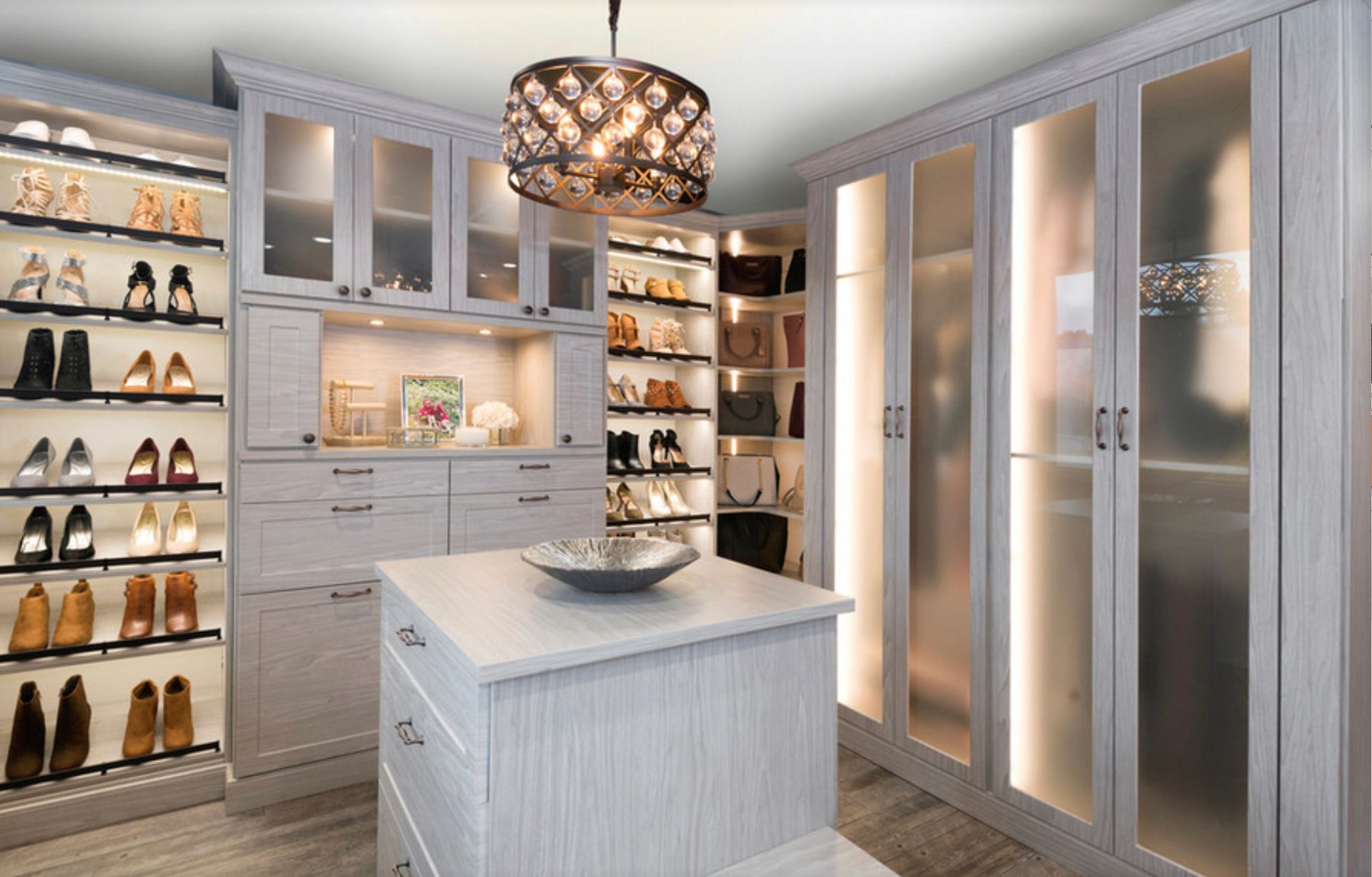 A walk-in closet with light wood cabinets, glass-door wardrobes, open shoe shelving, and a central island with a bowl.