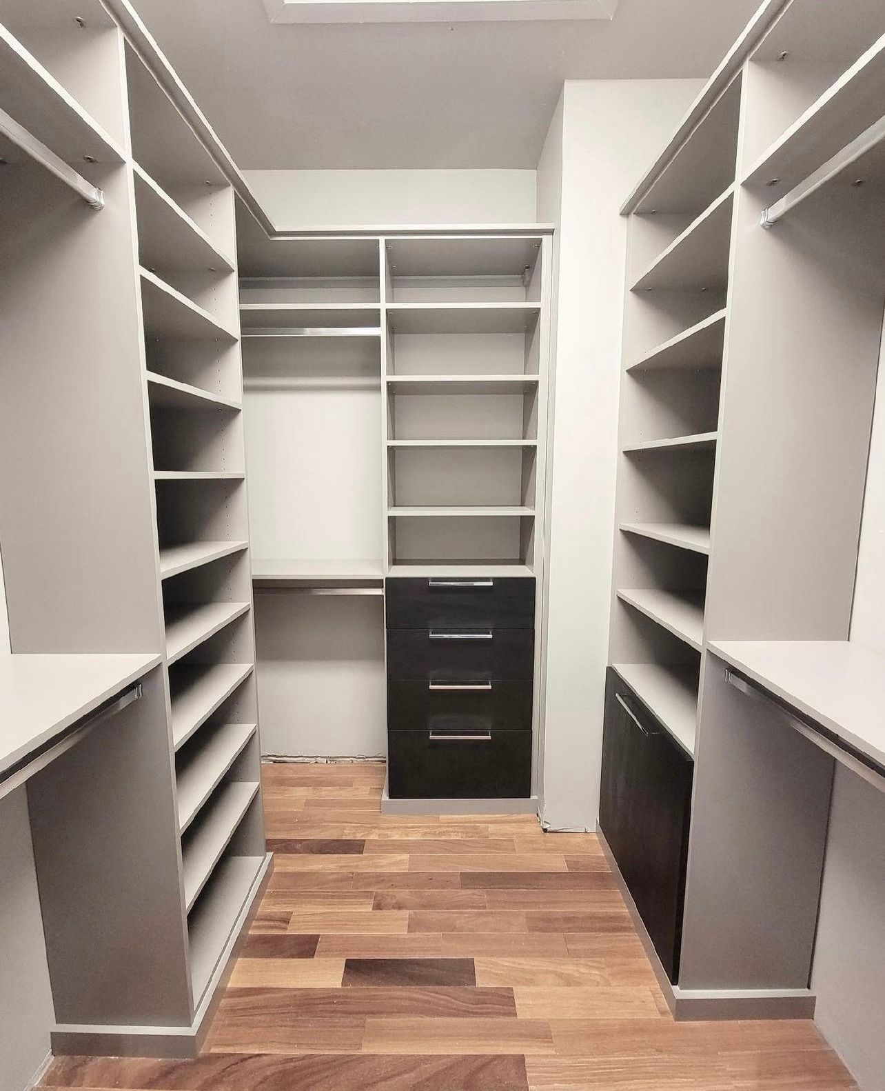 A walk-in closet featuring light gray shelving units, dark cabinet drawers, and hardwood flooring.
