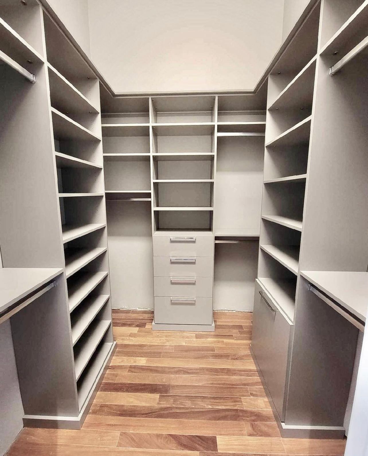 A walk-in closet with light grey custom shelving, built-in drawers, and wooden plank flooring.