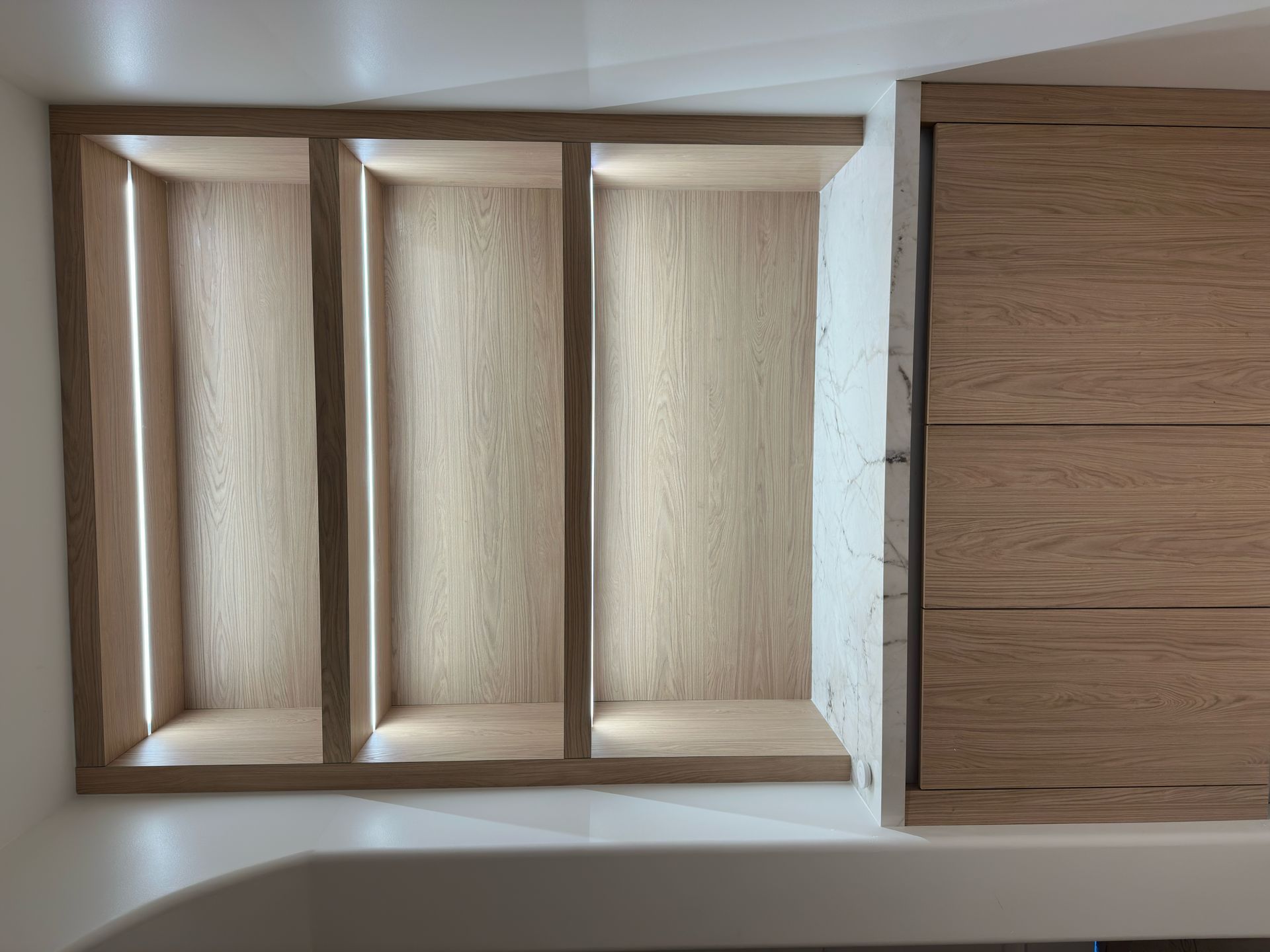 Three recessed, light wood shelves built into a wall next to wood-paneled cabinetry above a white bathroom fixture.