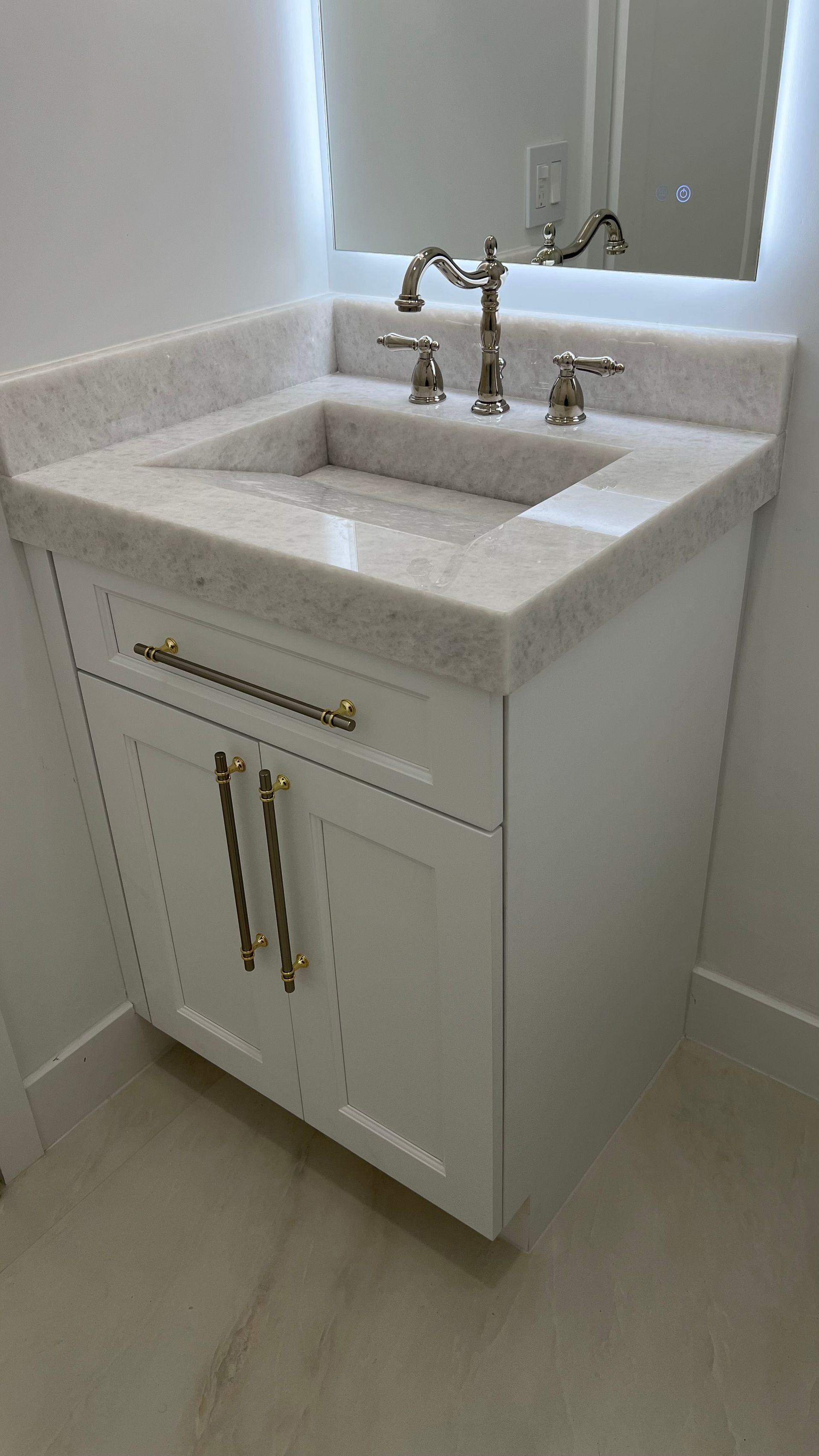 A white bathroom vanity with a speckled marble countertop, brass handles, and a modern gold faucet below a lit mirror.