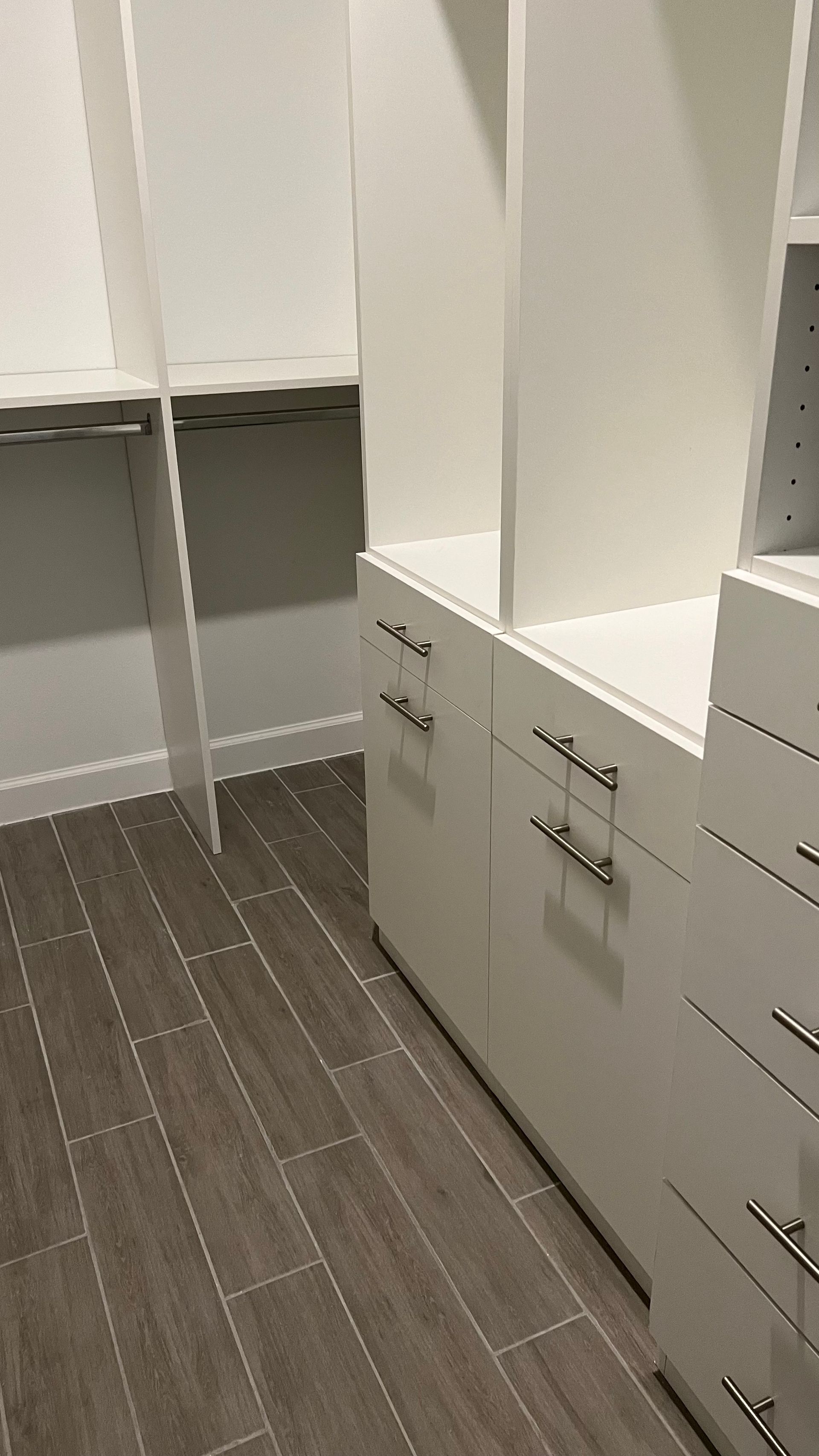 A custom white closet organizer with drawers and hanging rods above dark wood-look tile flooring.
