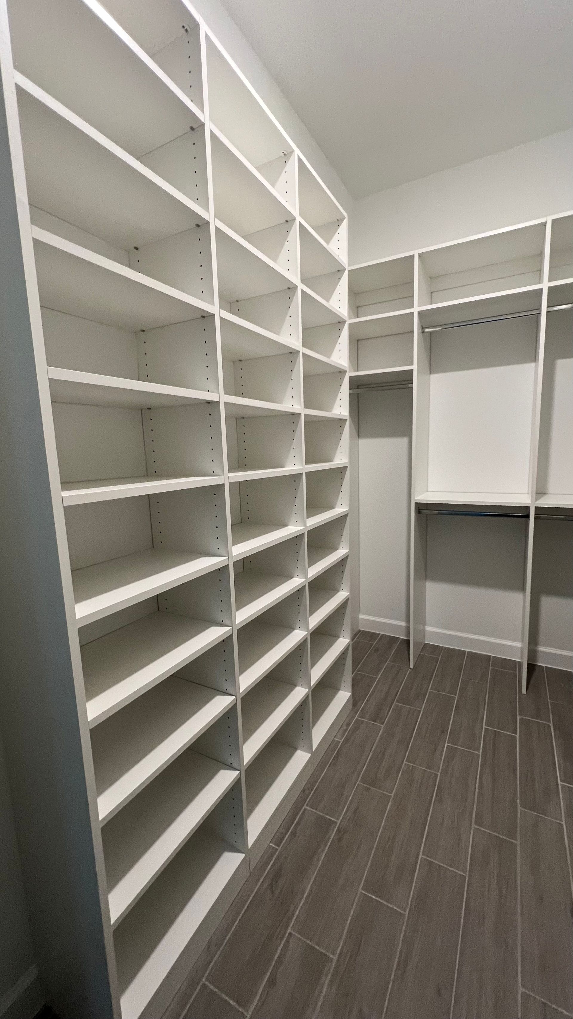 A walk-in closet featuring white floor-to-ceiling shelving units and a hanging rack against grey wood-look tiled floors.
