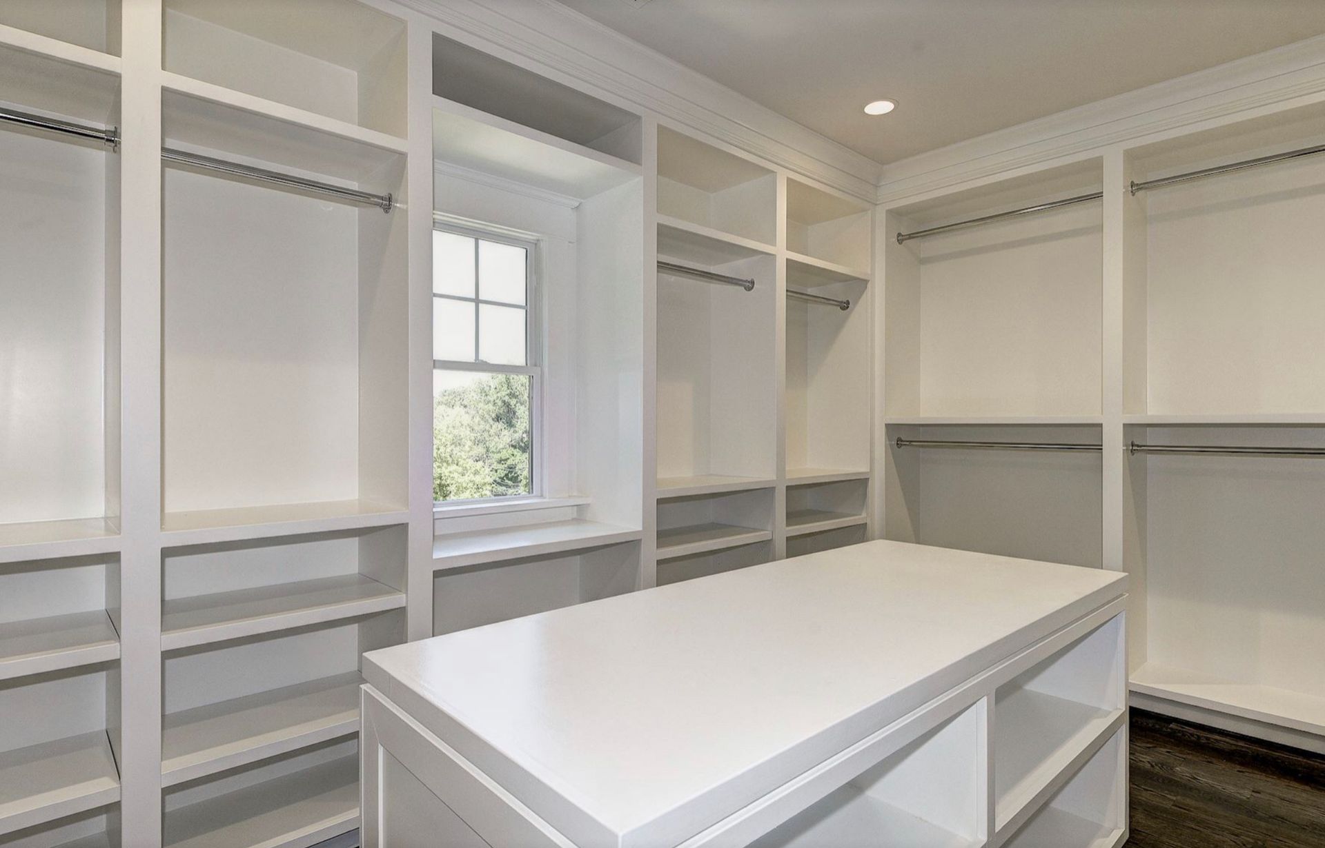 A walk-in closet featuring white wooden shelving, clothing rods, and a center island, lit by a small window.
