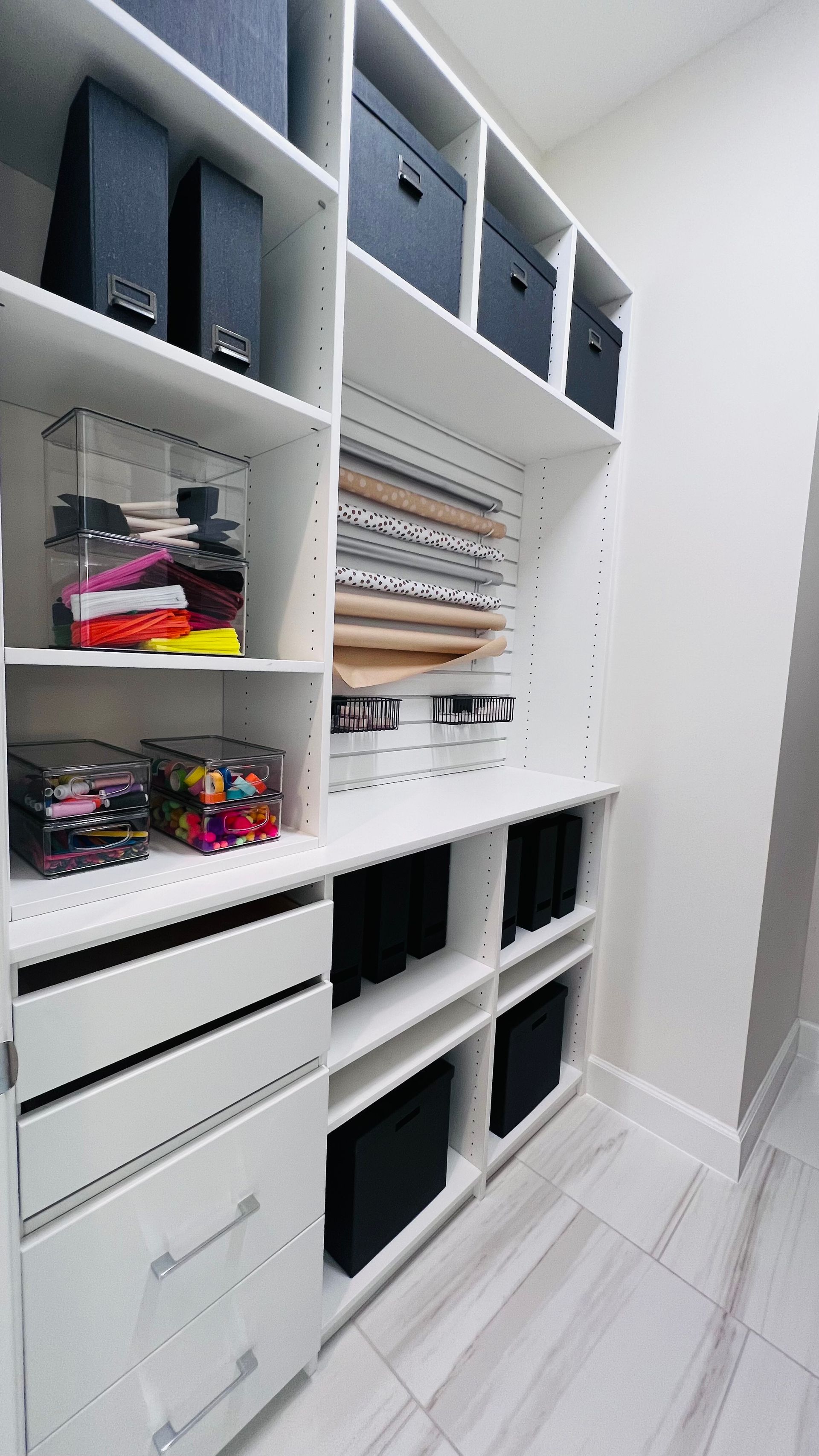 A white built-in storage unit with shelves, drawers, and black bins, used for organizing craft supplies and paper rolls.