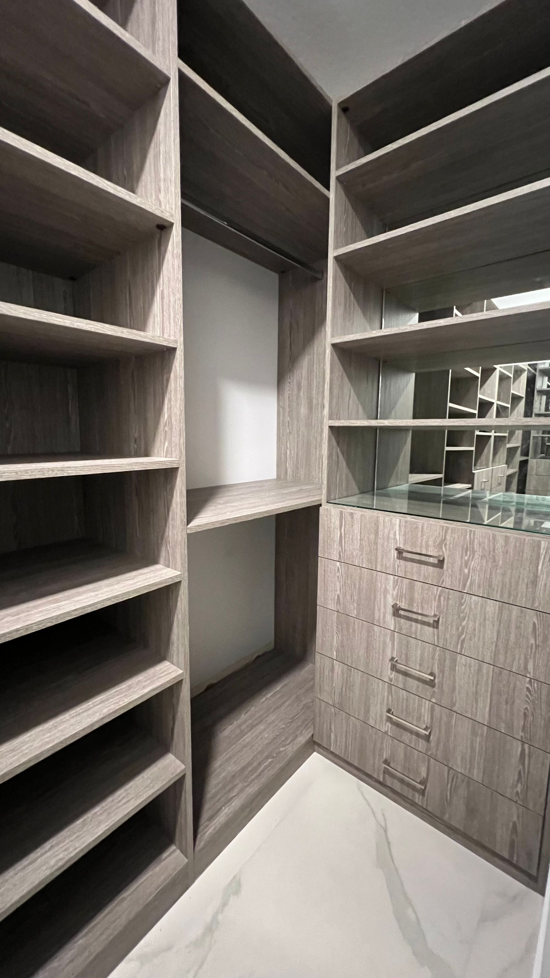 A modern walk-in closet with light wood shelving, a central hanging area, and a column of five drawers with metallic pulls.