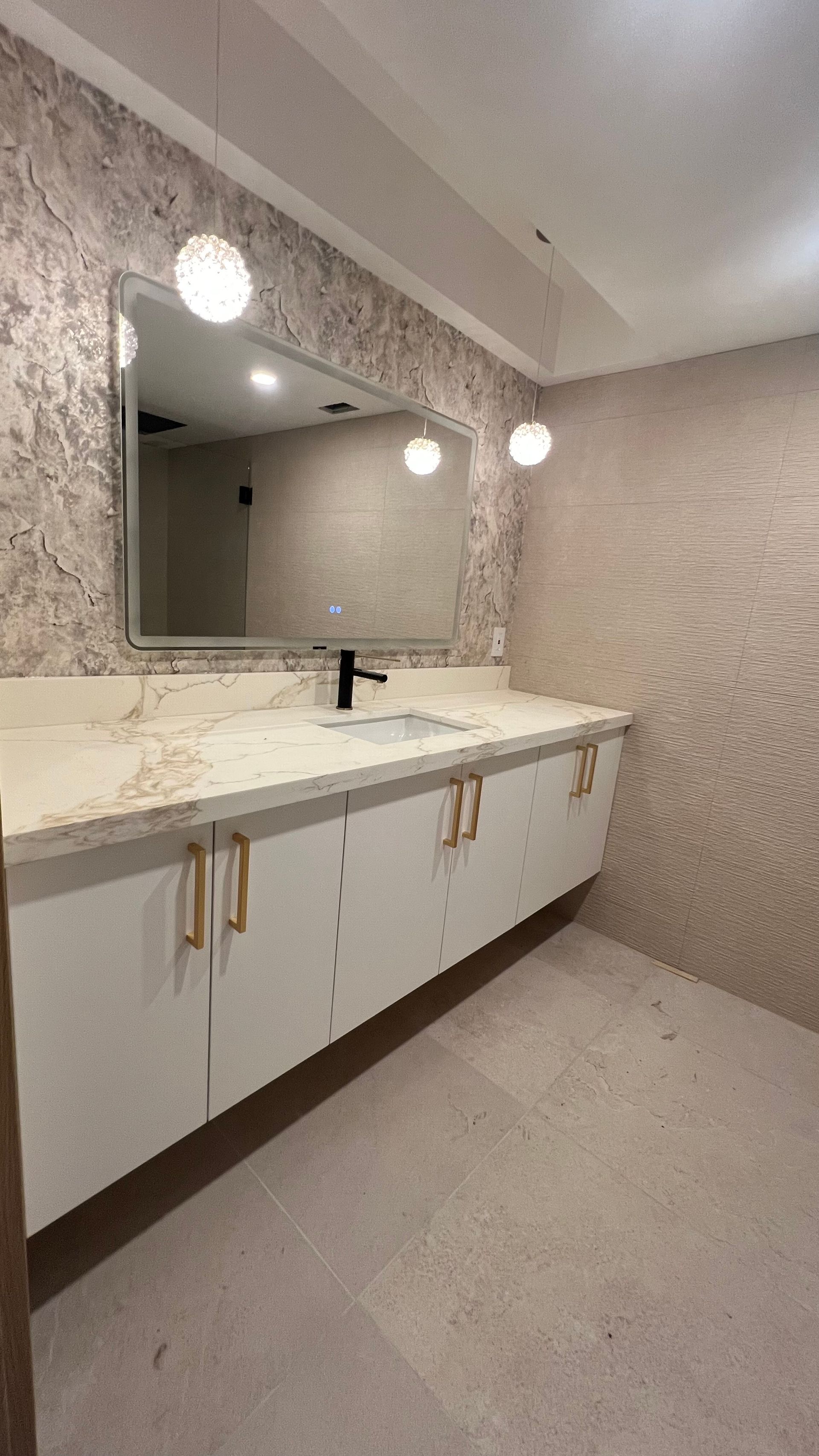 Modern bathroom vanity with a marble countertop, white cabinets with gold handles, a wall mirror, and textured wallpaper.