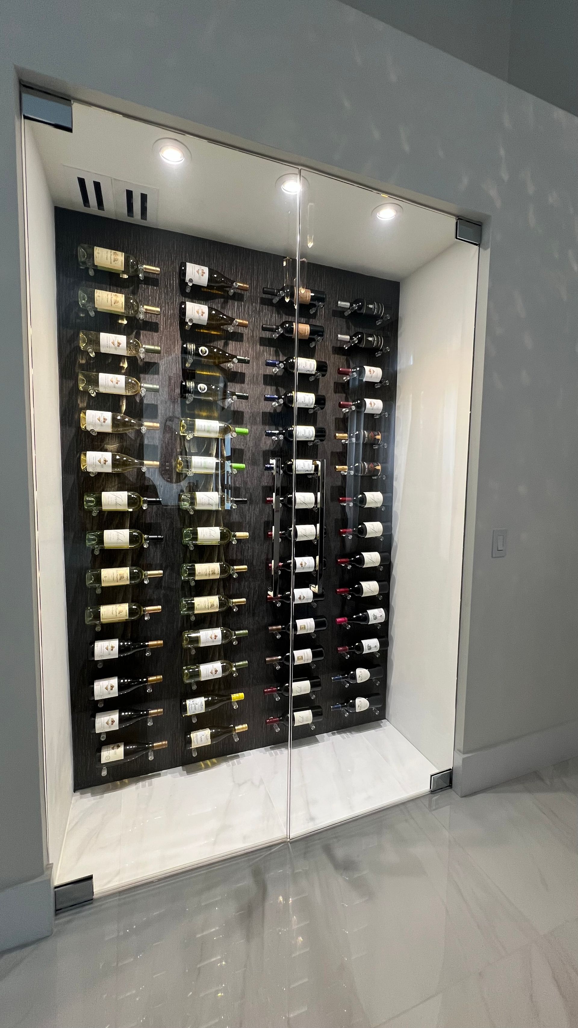 A modern, recessed glass-fronted wine cellar with illuminated shelving holding multiple bottles against a black wall.