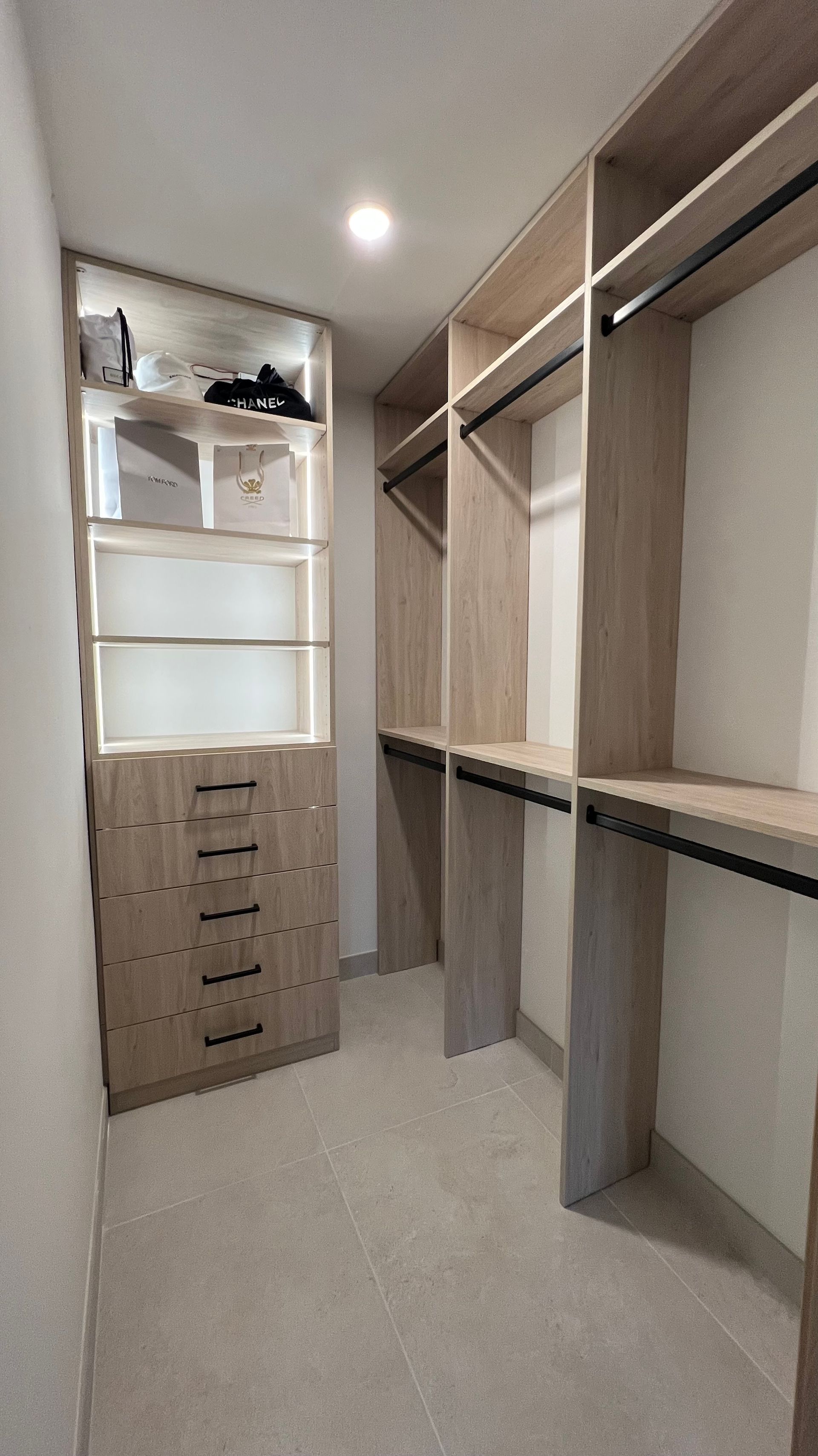 A walk-in closet with light wood shelving, a central stack of five drawers, and metal clothing rods on the side walls.