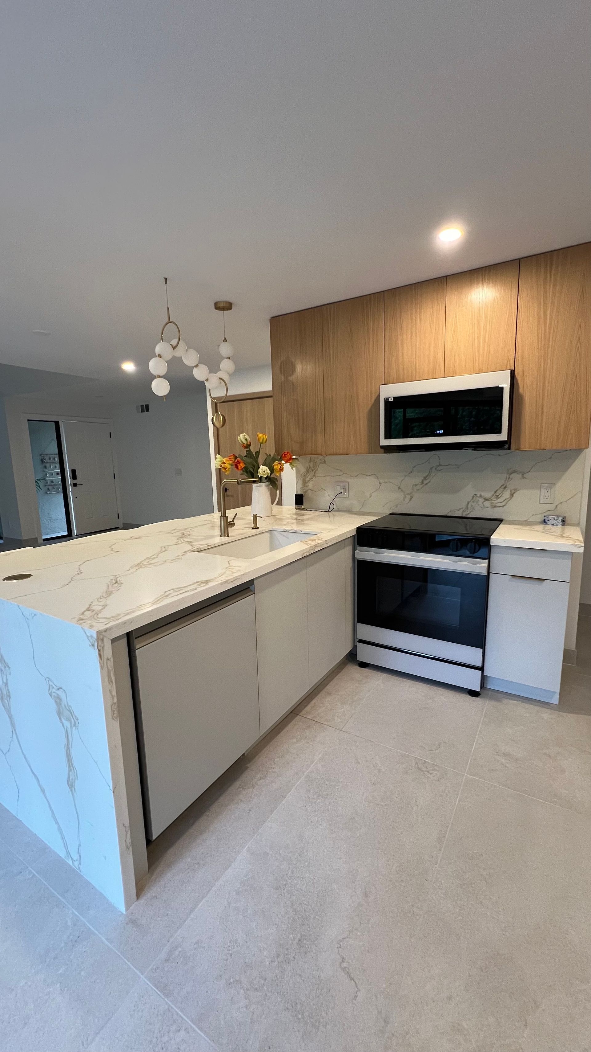 A bright modern kitchen with a white marble island, light wood cabinets, stainless steel appliances, and tile flooring.
