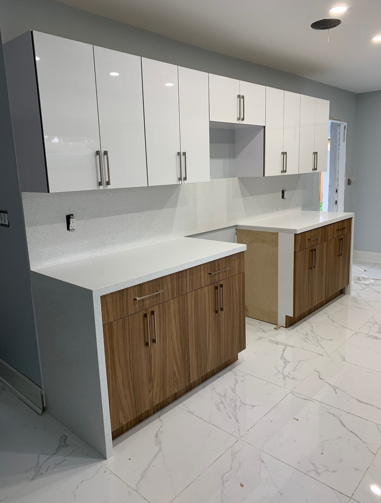 A kitchen under renovation with white upper cabinets, wood-grain lower cabinets, white quartz countertops, and tile floors.