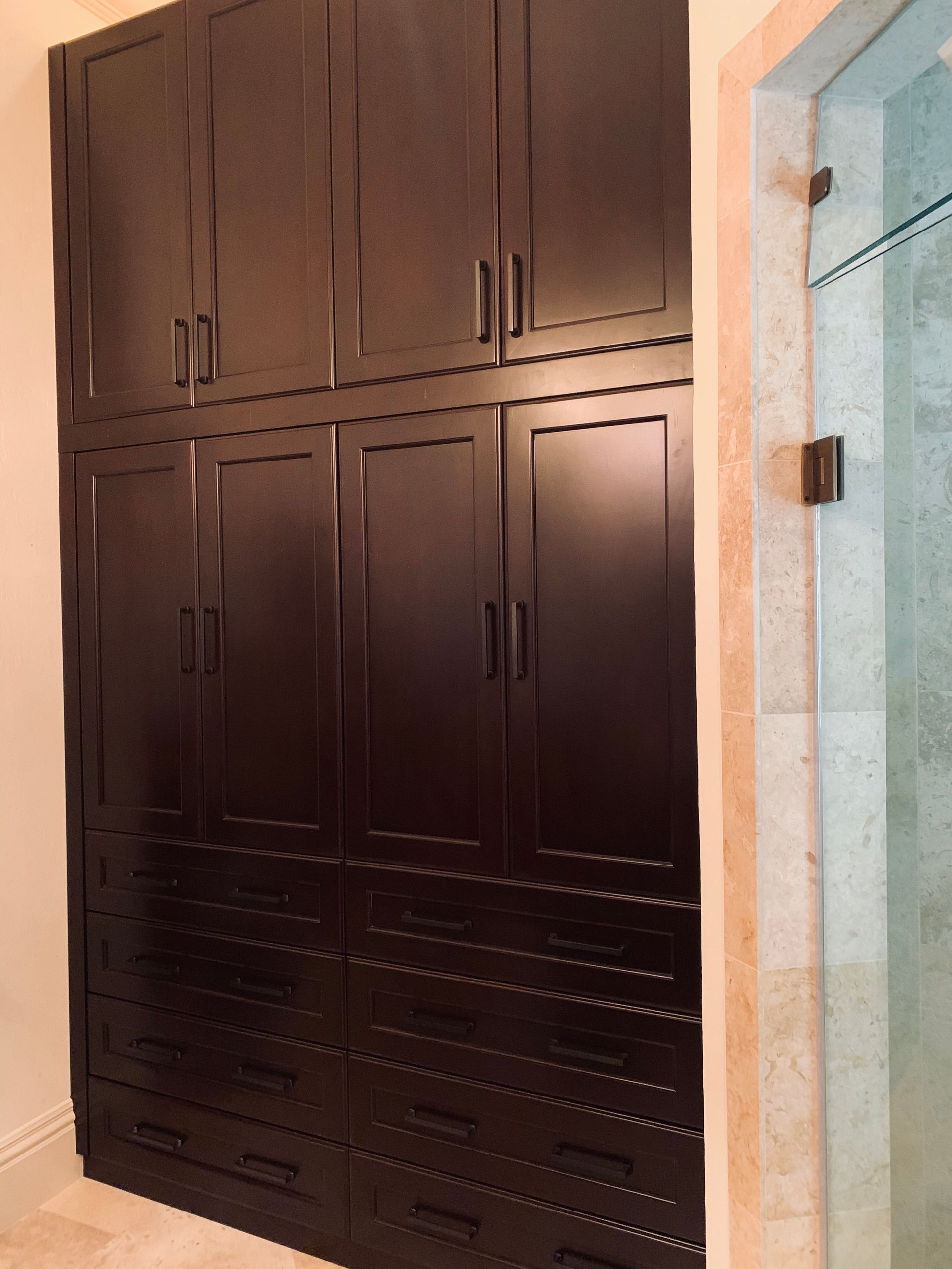 A large, floor-to-ceiling dark brown wooden storage cabinet with multiple doors and drawers, next to a glass shower.
