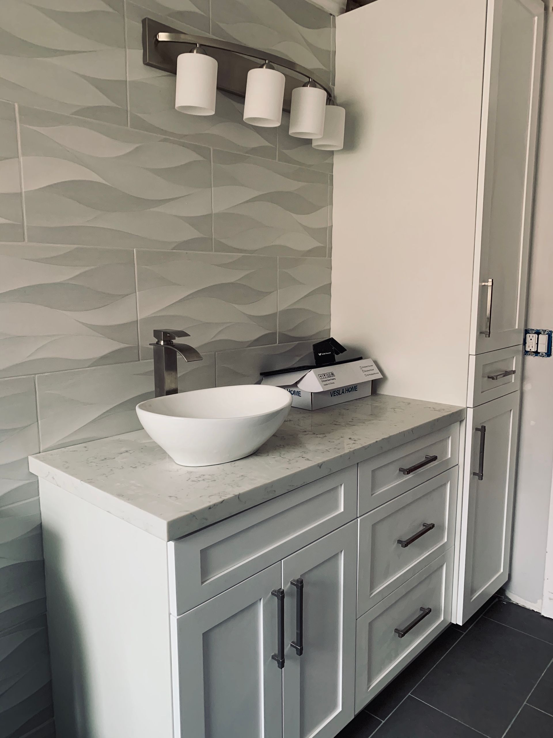 Modern bathroom vanity with a white vessel sink, quartz countertop, and grey wavy-patterned tiled wall.