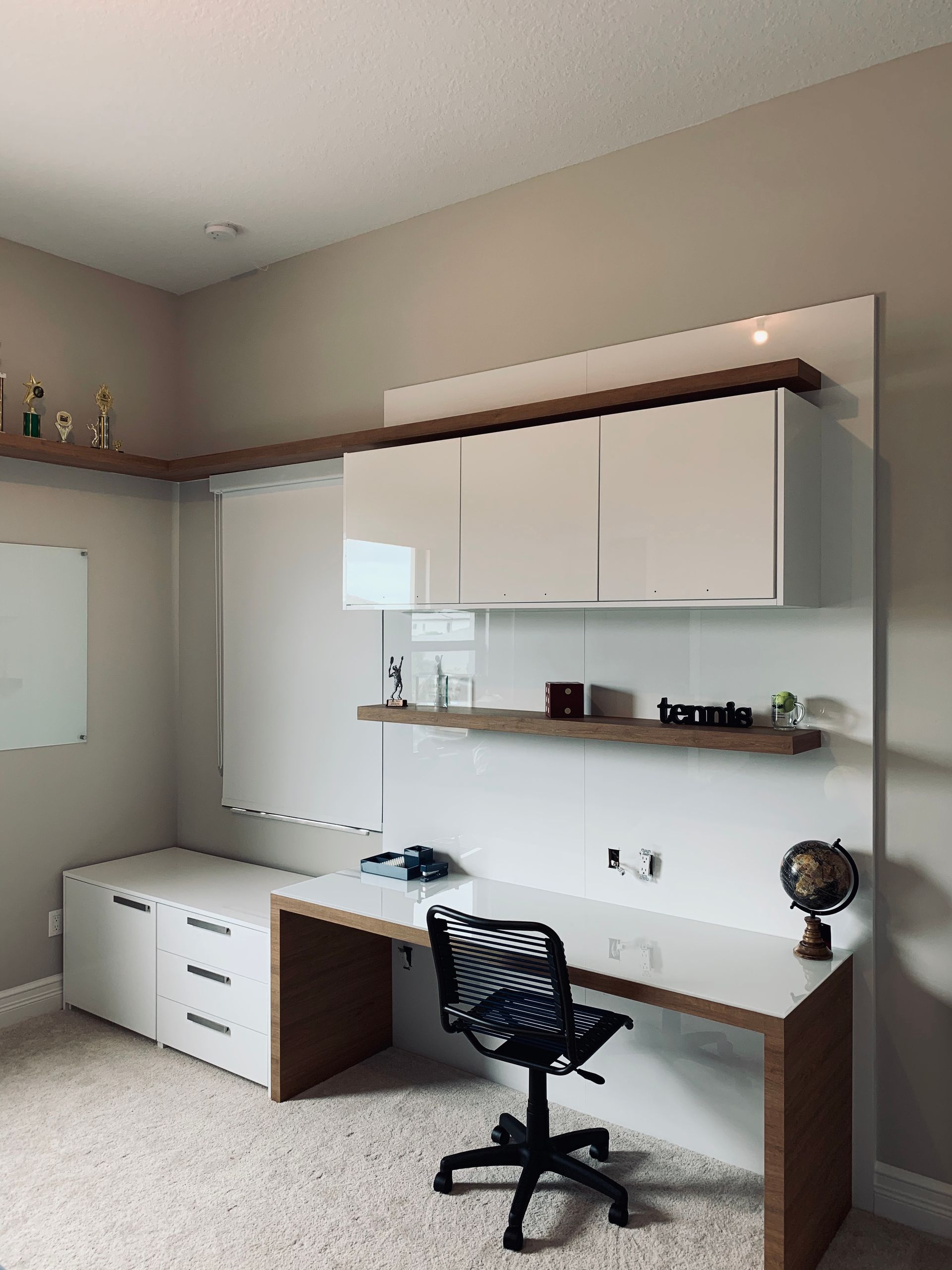 A modern home office with a white desk, matching cabinets, wood shelving, and a black mesh office chair on a carpet floor.