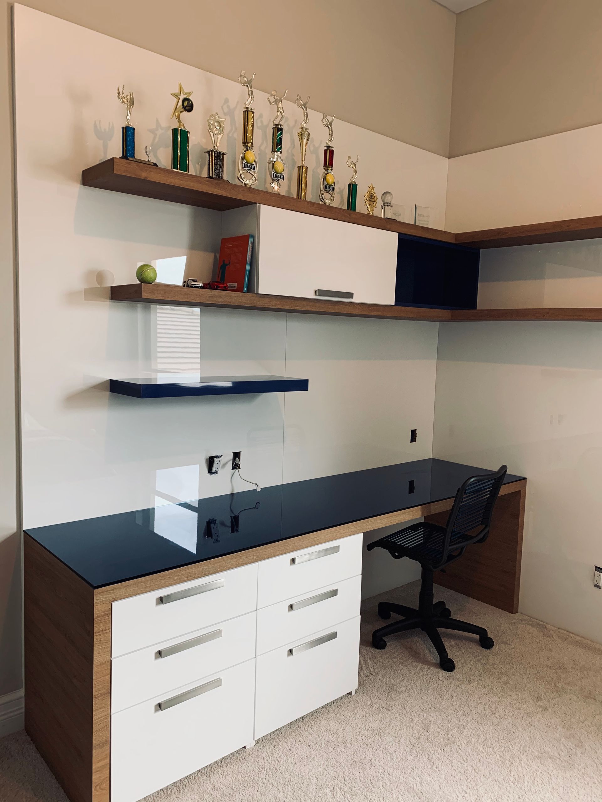 A modern office desk with white drawers, a black top, and floating wood shelves displaying trophies against a beige wall.