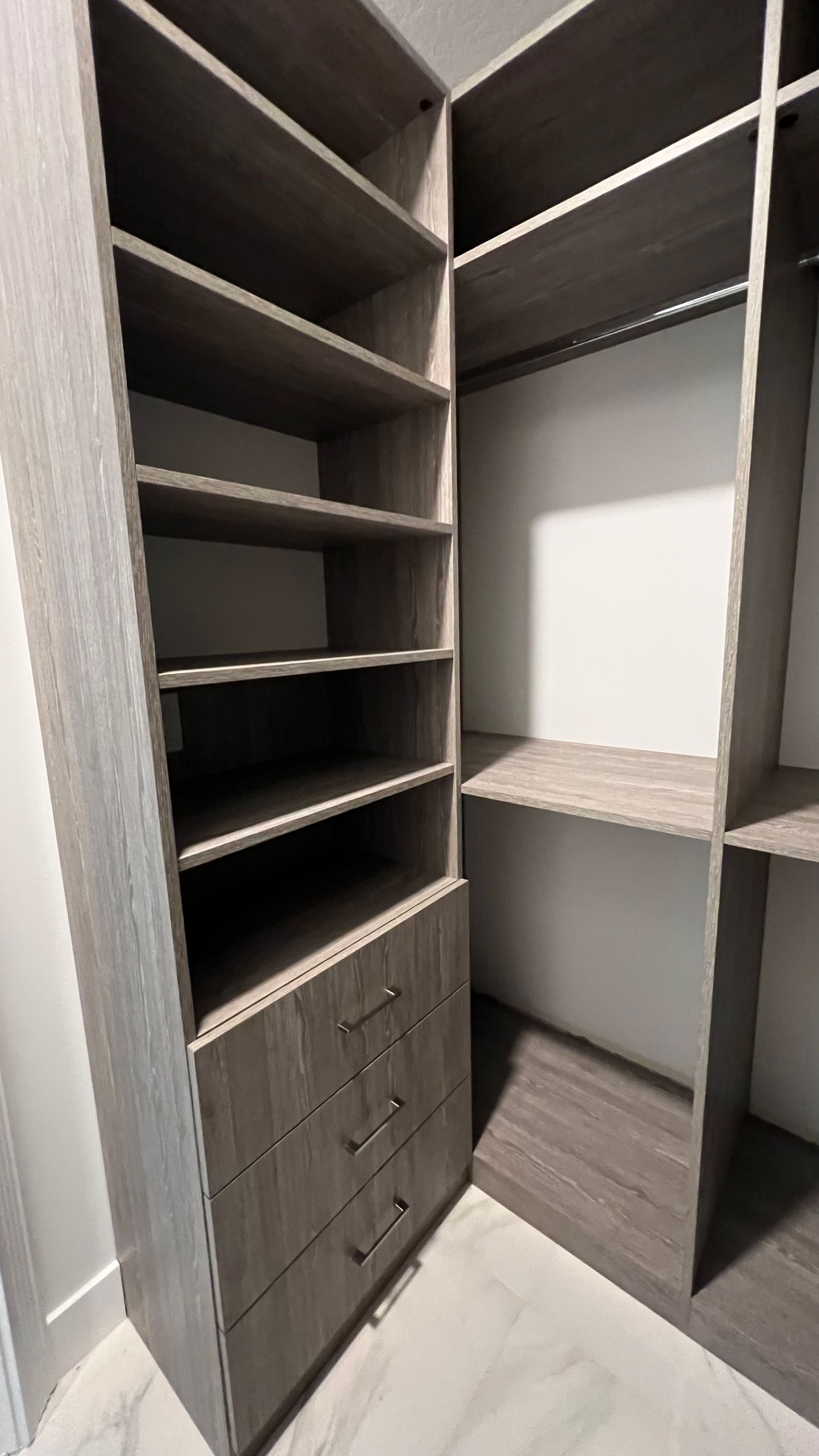 Modern walk-in closet featuring grey wood-grain shelving, hanging rods, and a three-drawer unit on white marble-look floors.