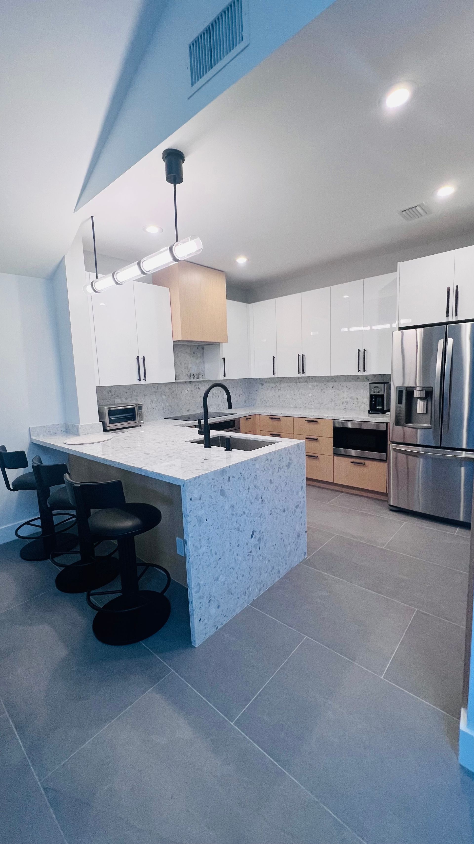 Modern kitchen with a white marble-topped island, light wood lower cabinets, white upper cabinets, and dark tile flooring.