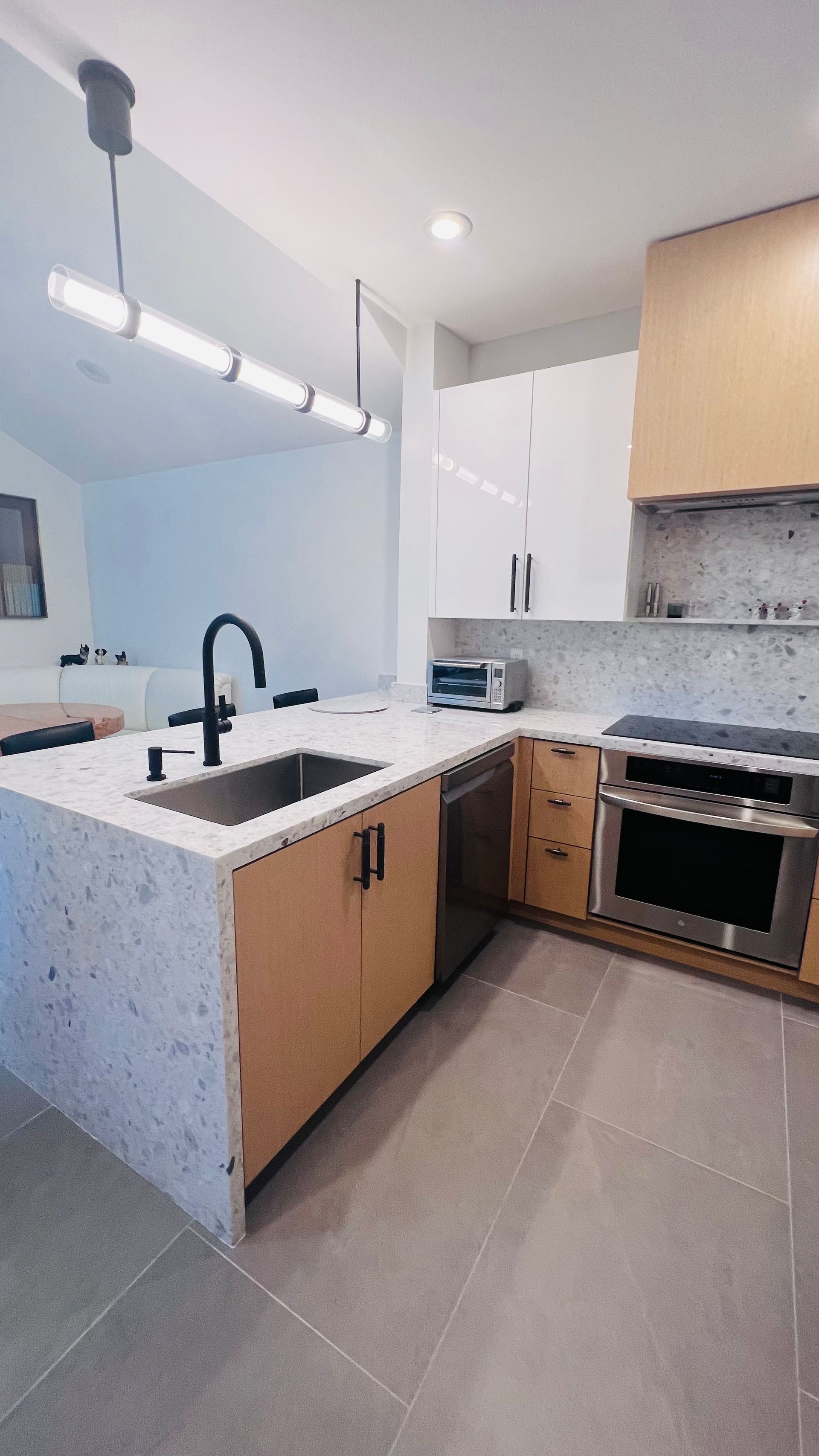 A modern kitchen with a stone-topped island, wooden cabinetry, a stainless steel stove, and a linear pendant light.