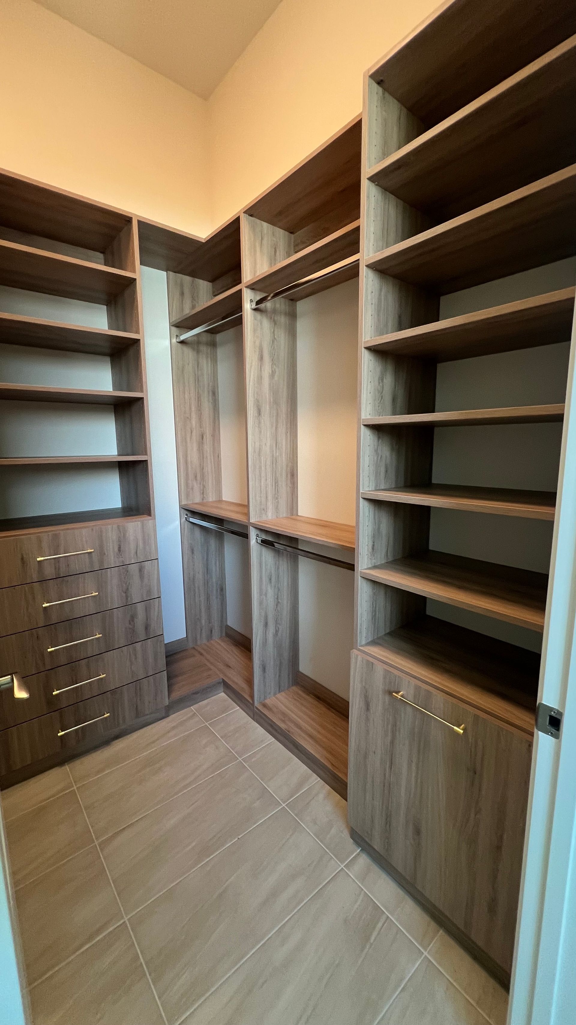 A walk-in closet featuring light brown wood-grain shelving, hanging rods, and a six-drawer cabinet on a tiled floor.