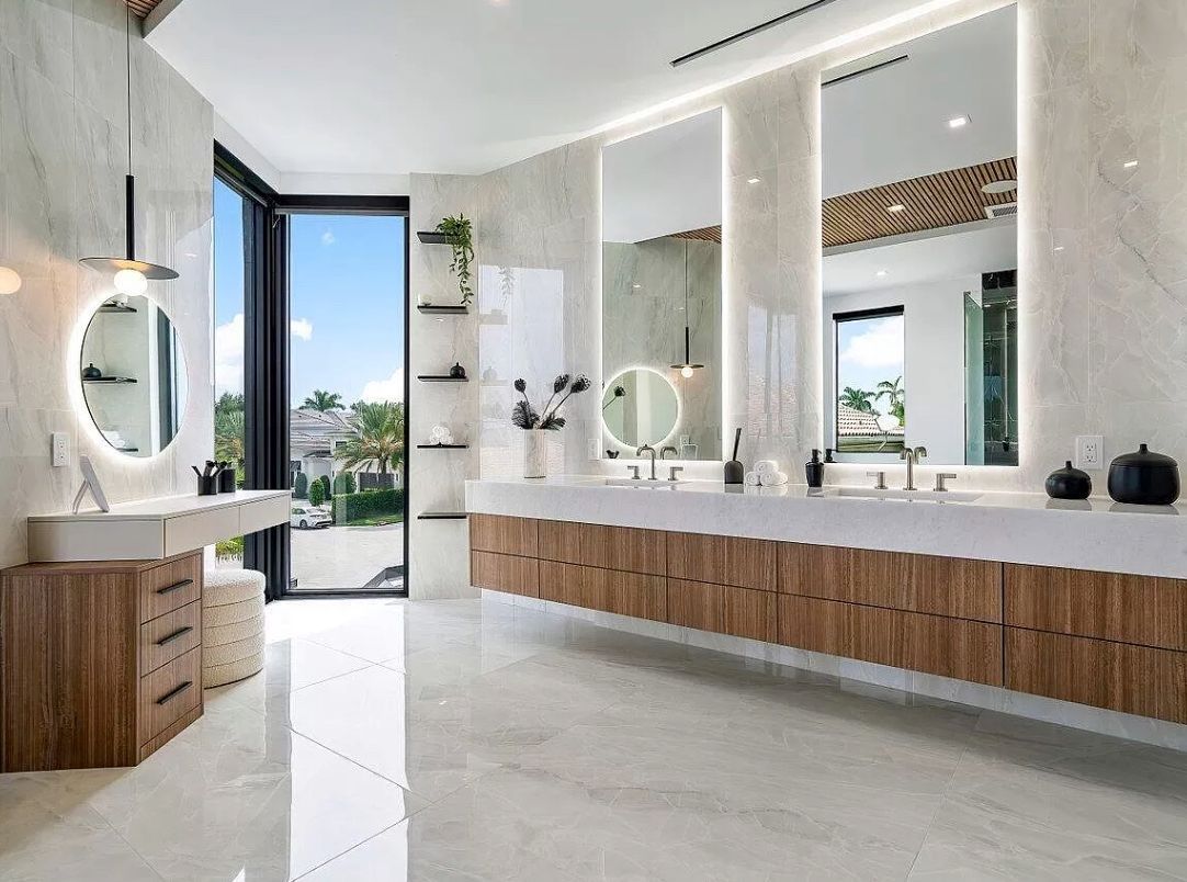 A modern bathroom with marble walls, wood cabinetry, dual sinks, mirrors, and a glass door leading to an outdoor view.