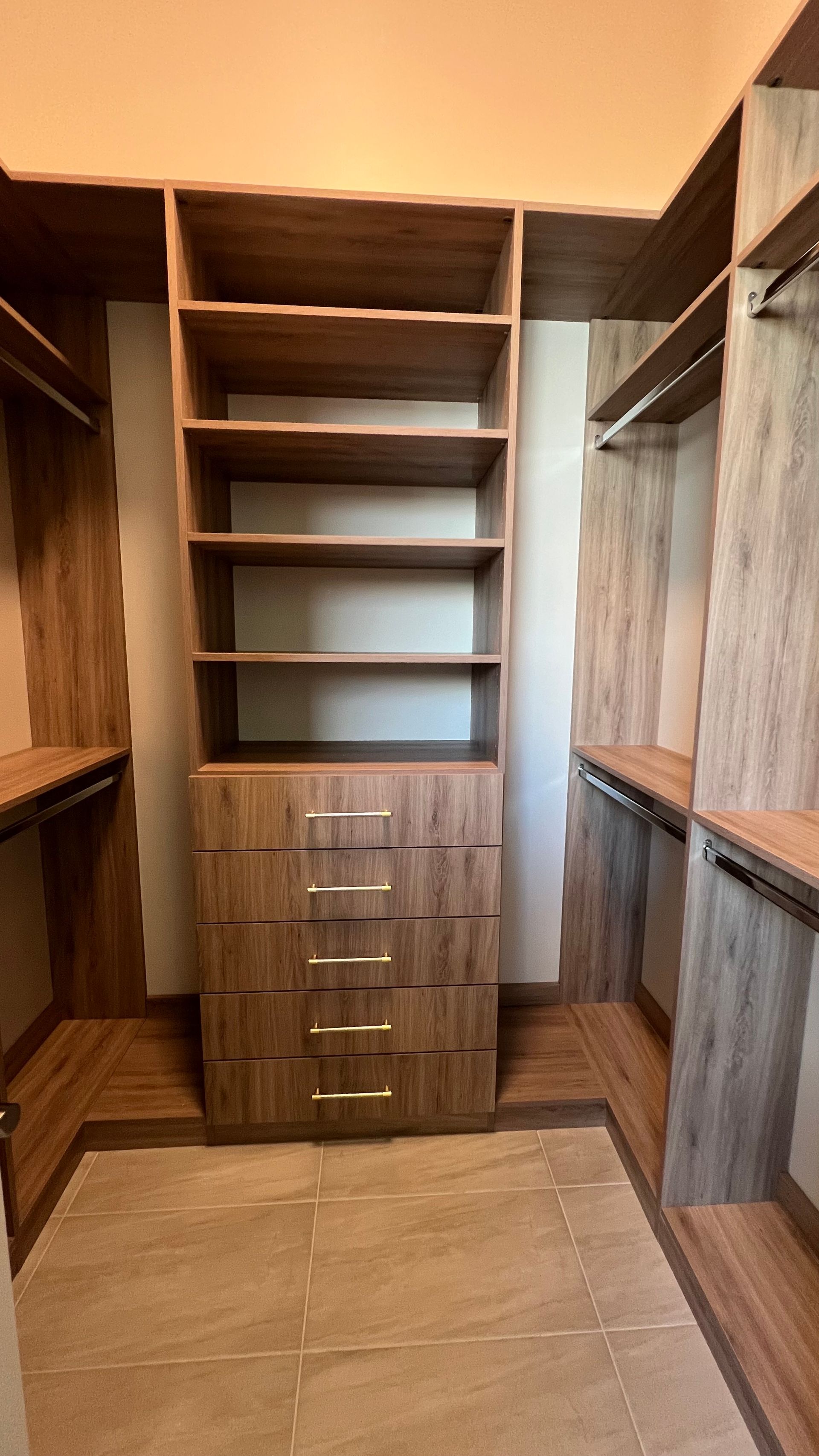 Walk-in closet with wood-grain finish, open shelving, a central five-drawer stack, and hanging rods on both sides.